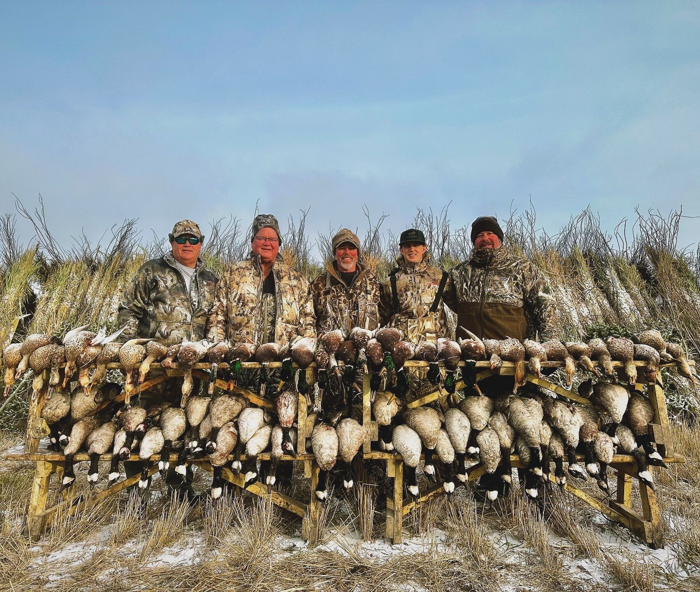 Five hunters posing behind a wall of harvested birds in a snowy field under a blue sky