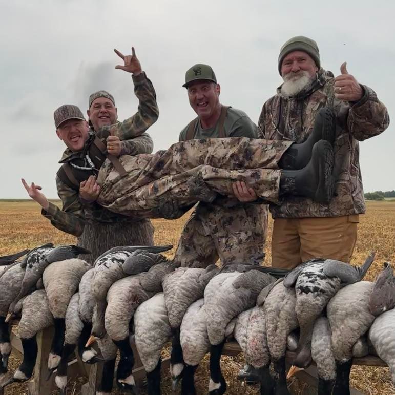 Four hunters posing behind a row of geese in a field, wearing camouflage and smiling