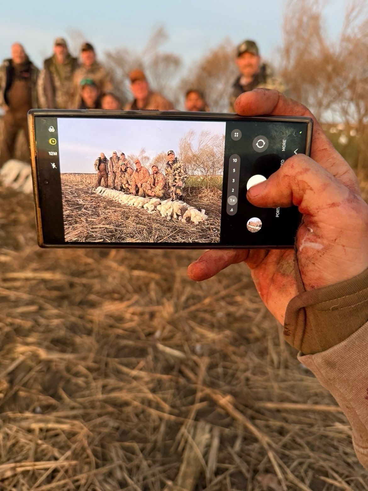 Hand holding a smartphone camera, photographing a group standing in a dry field at sunset