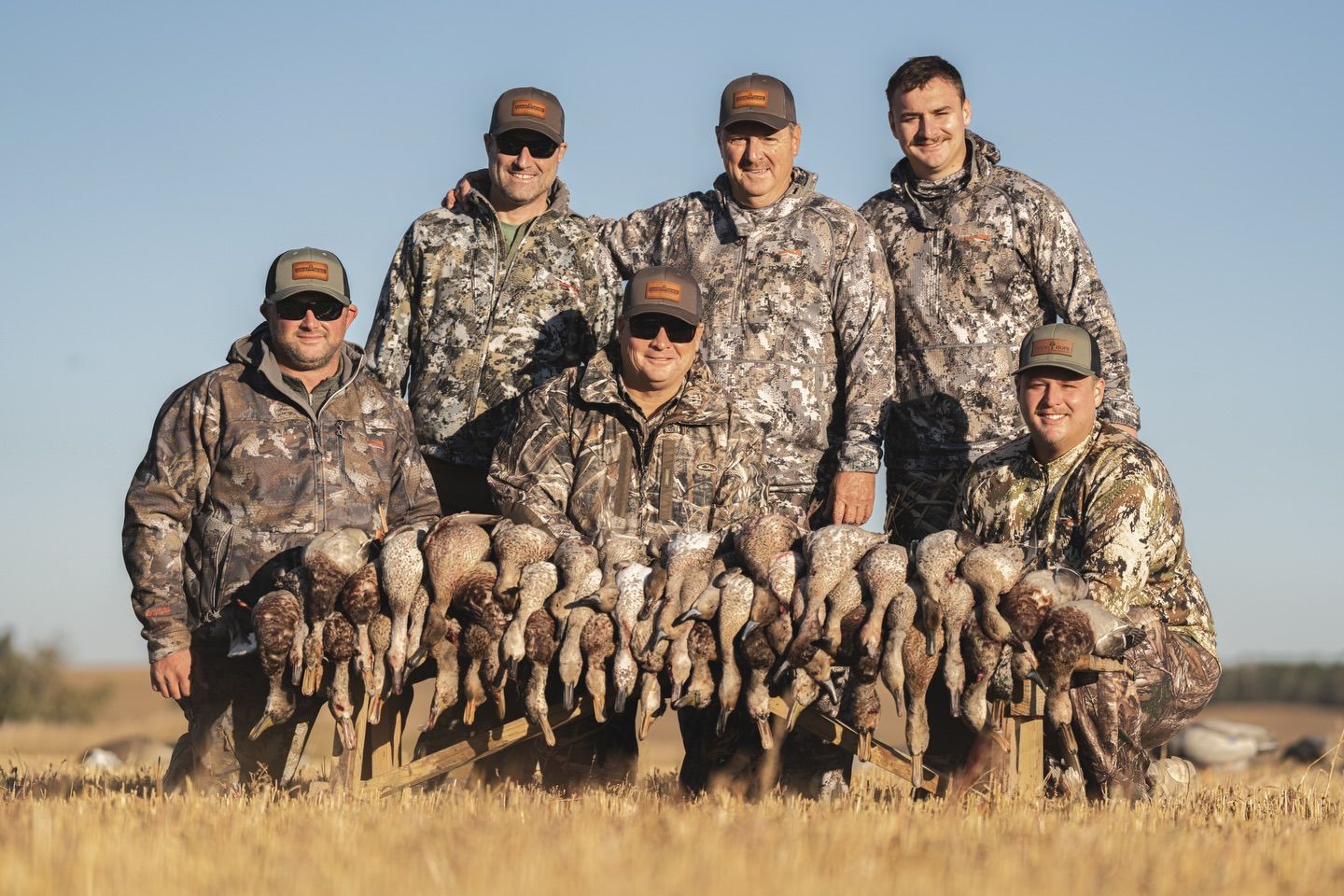 Six hunters in camouflage pose with a row of harvested ducks in a grassy field.