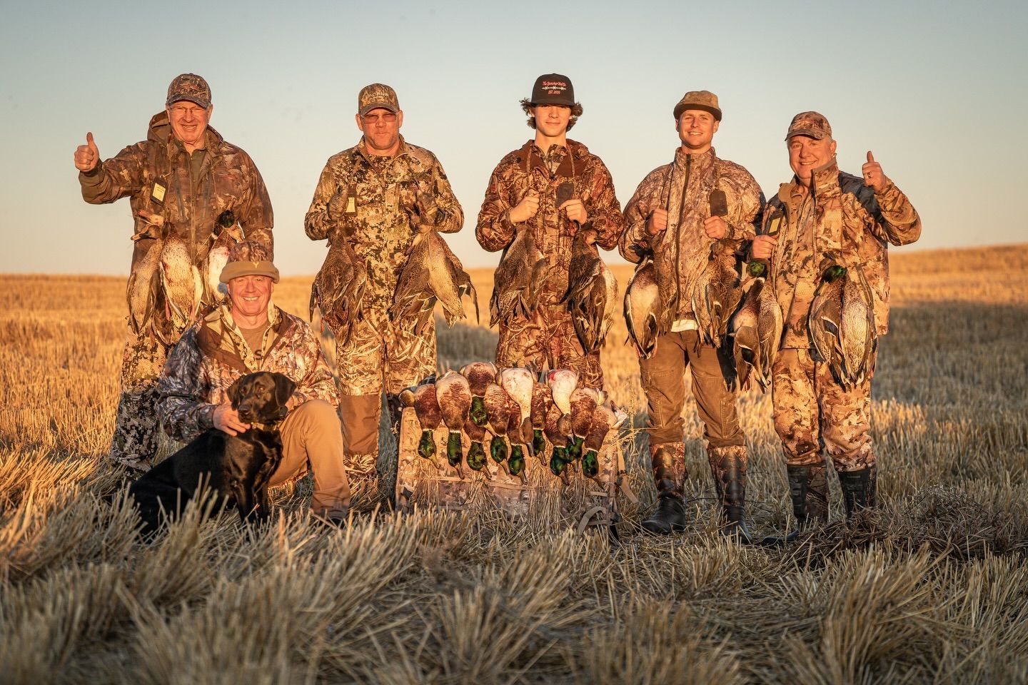 Seven hunters in camouflage pose with two dogs in a grassy field at sunset.