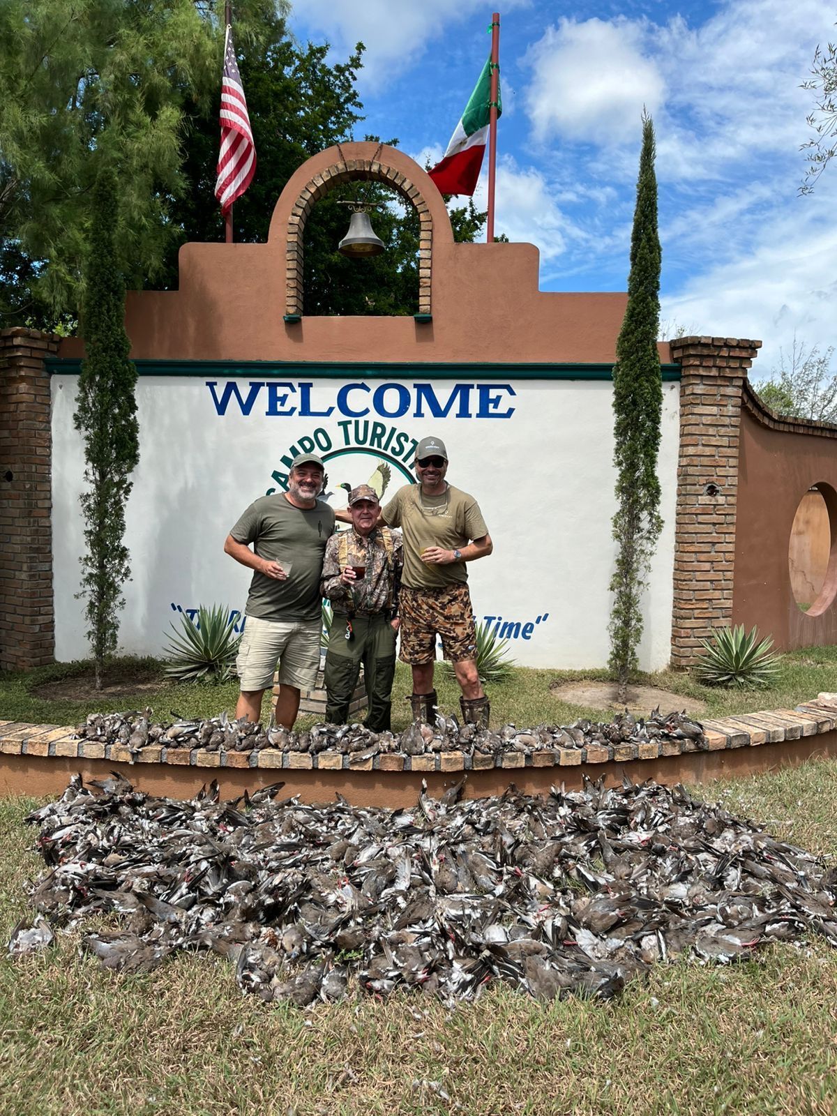 Two soldiers posing at a Fort Hood welcome sign with flags and cactus plants behind them.