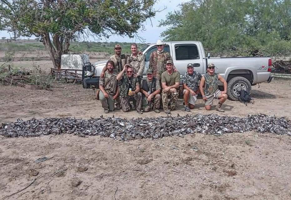 Group of people in camouflage posing beside a pickup truck in a dry, rural field with a pile of wild boar