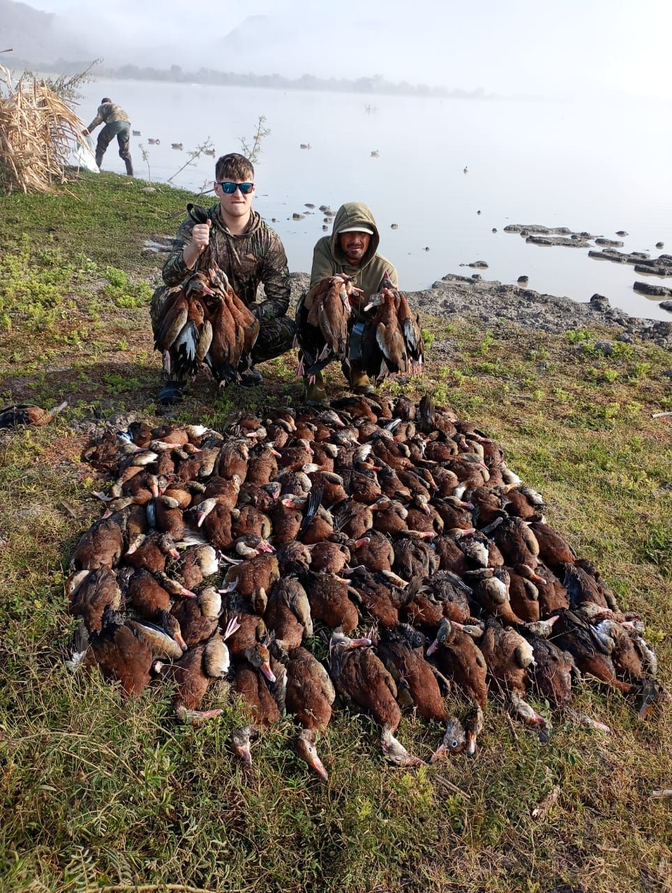 Two hunters kneel beside a large pile of ducks by a lakeshore in cold, foggy light