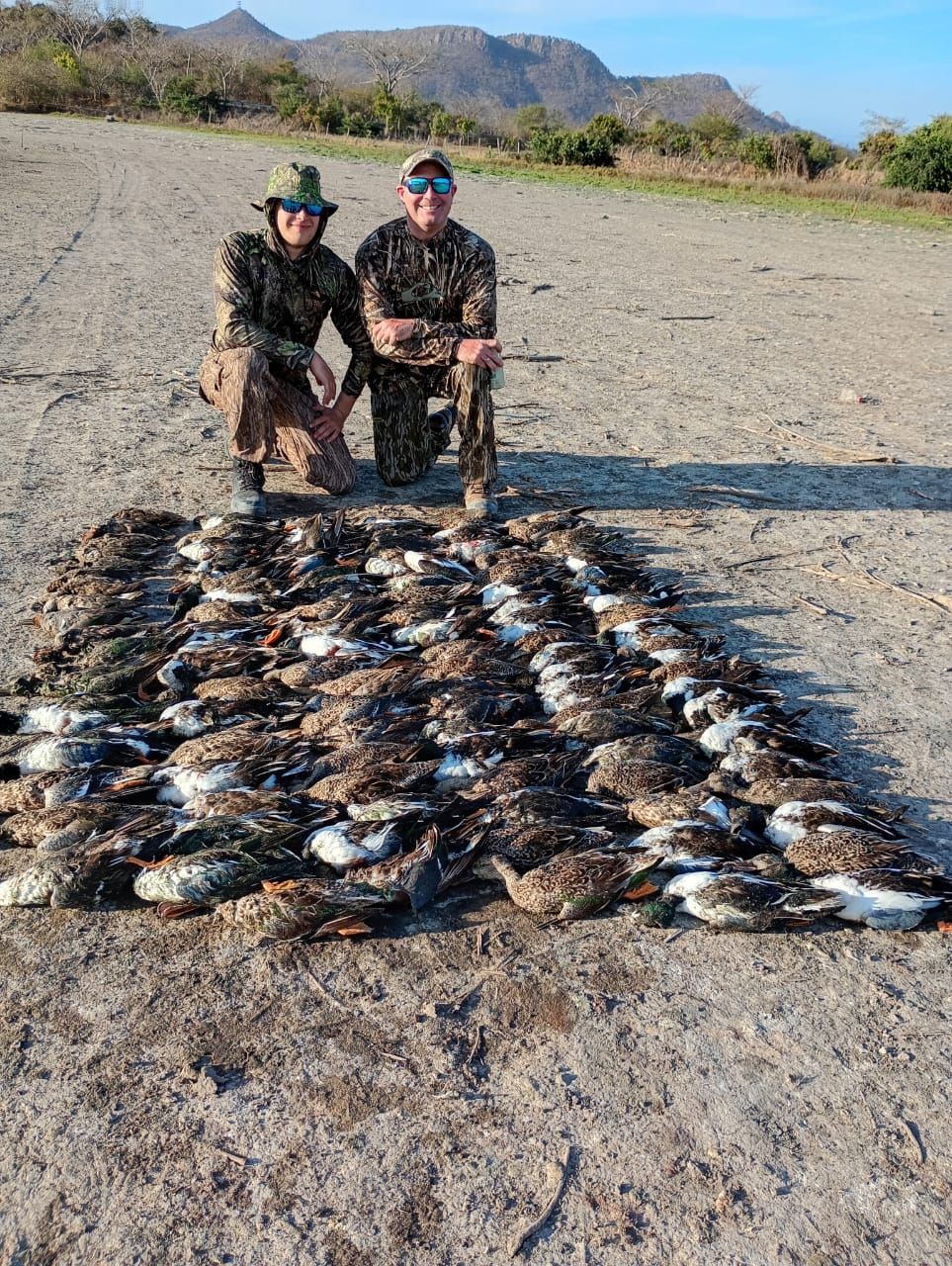 Two hunters kneel behind a large spread of dead birds on a rocky desert plain.