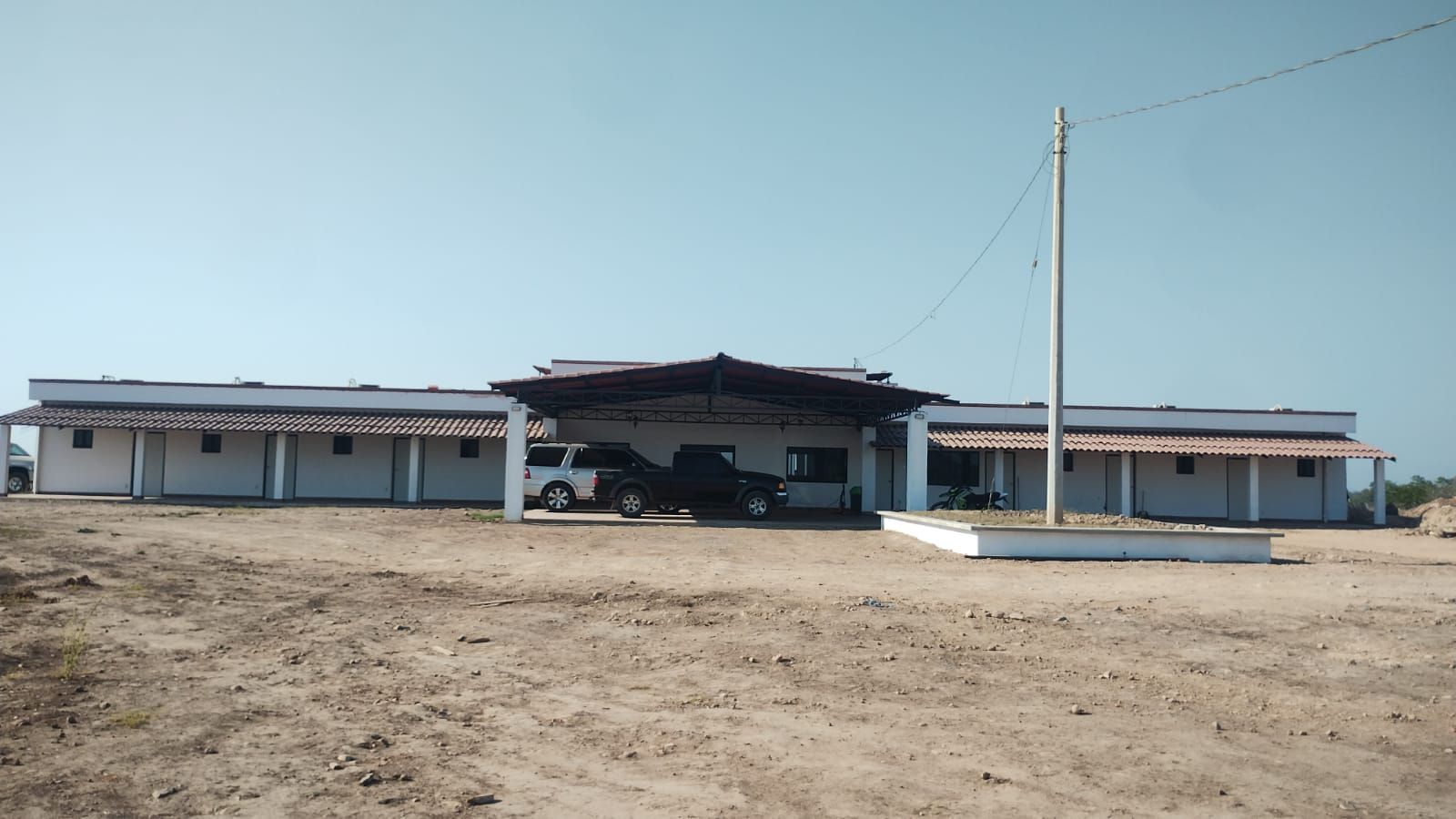 A low, white building in a dry field under a clear blue sky, with a tall utility pole in front.