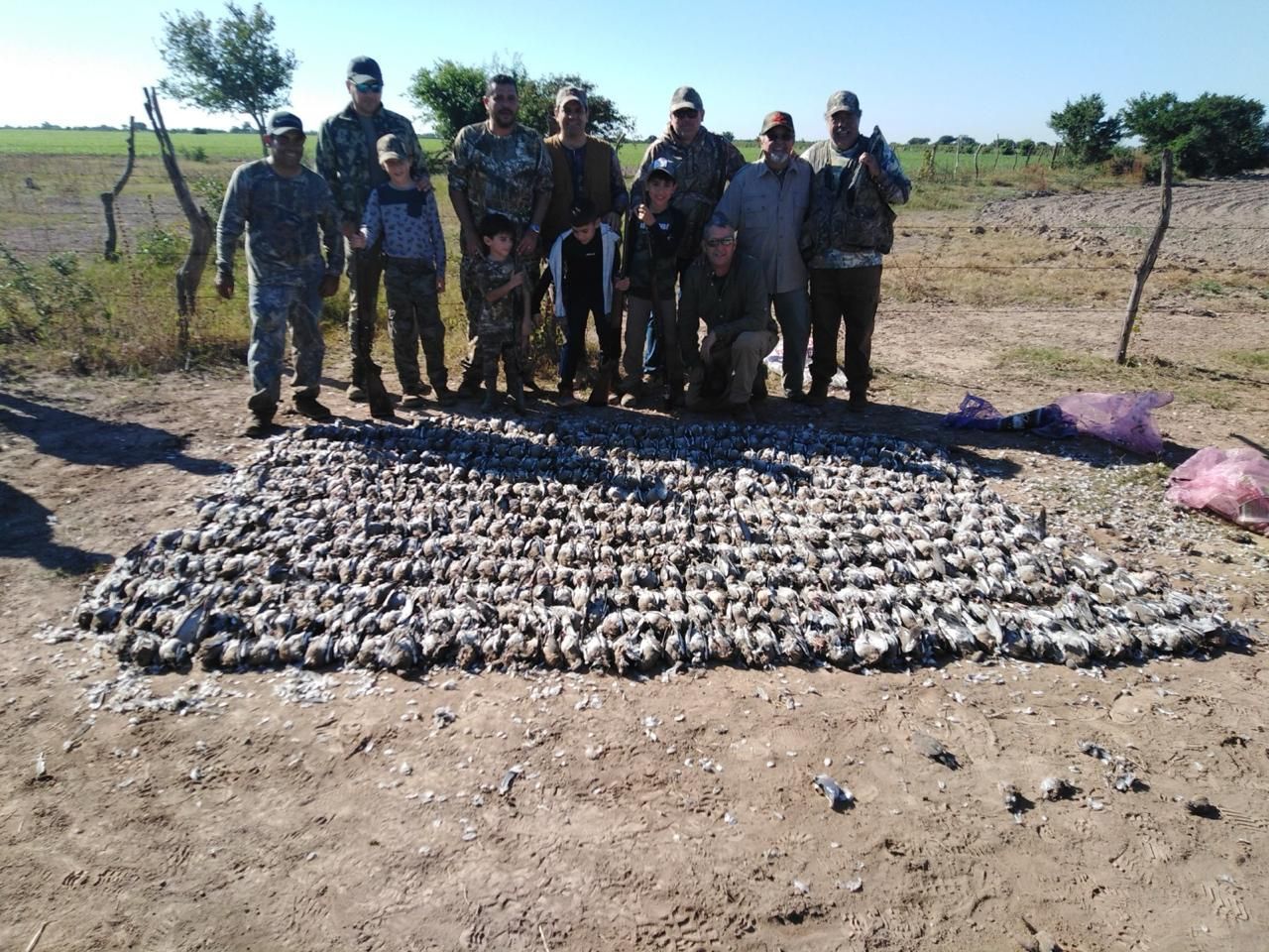 Group of people standing behind a large pile of harvested game in a dry field