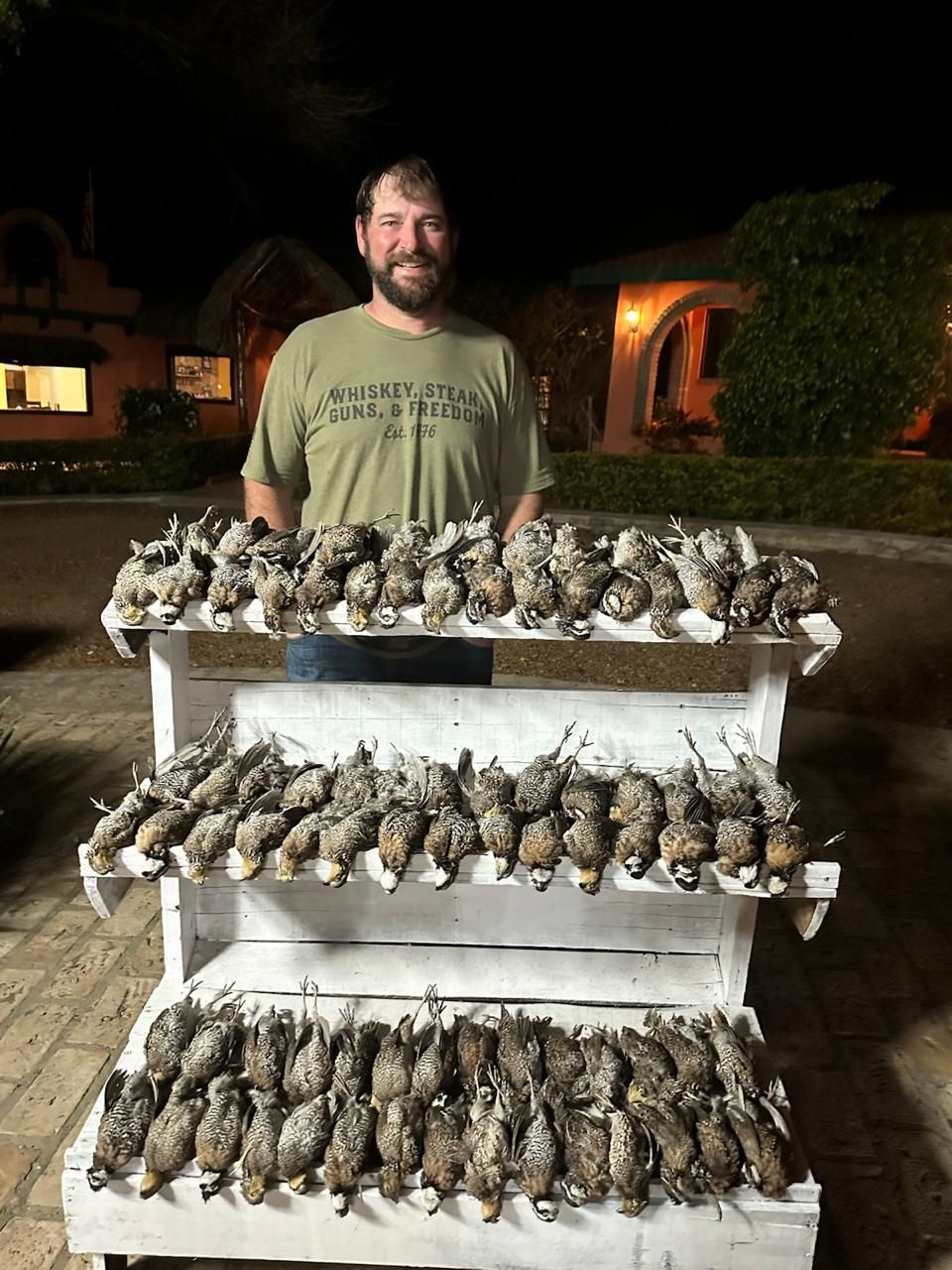 Man standing behind a table covered with rows of dead fish or birds at night outdoors