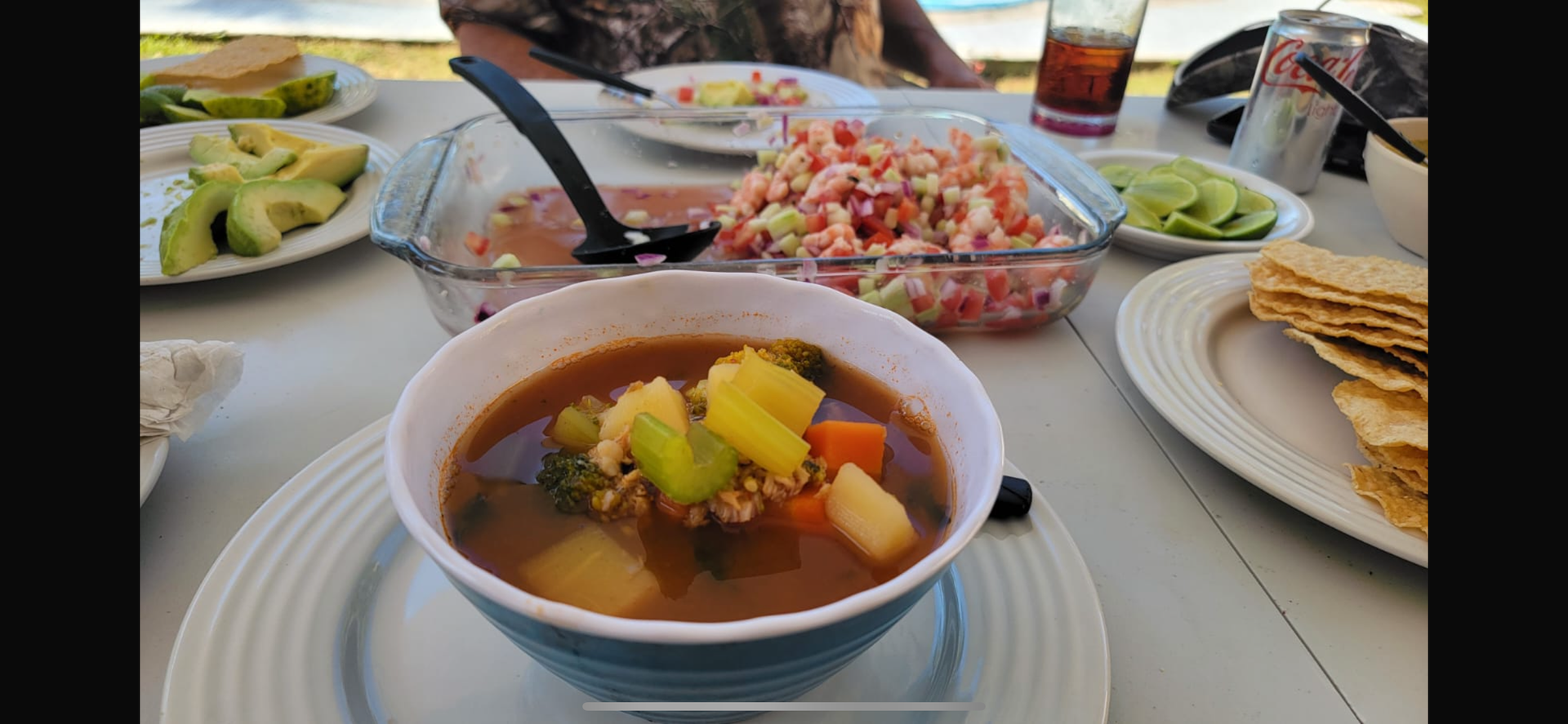 Outdoor table with bowls of soup, salad, avocado slices, and crackers in a bright dining setting