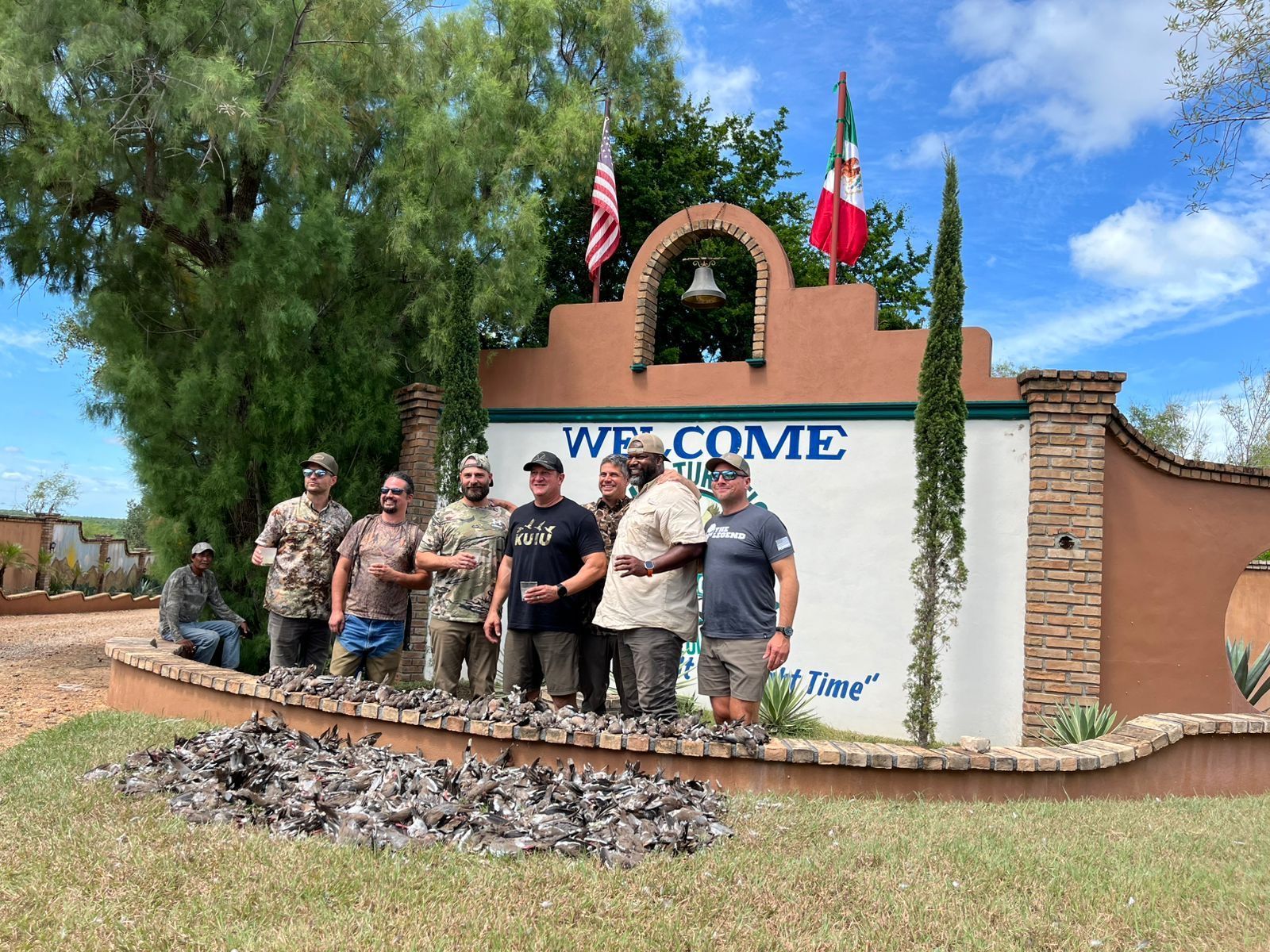 Six people posing in front of a stone-and-stucco welcome sign with flags and greenery outdoors