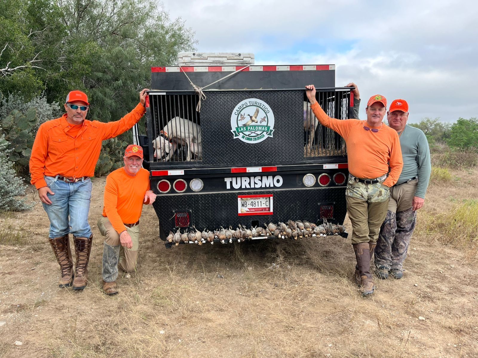 Four people in orange gear pose beside a black turismo truck in a grassy field.