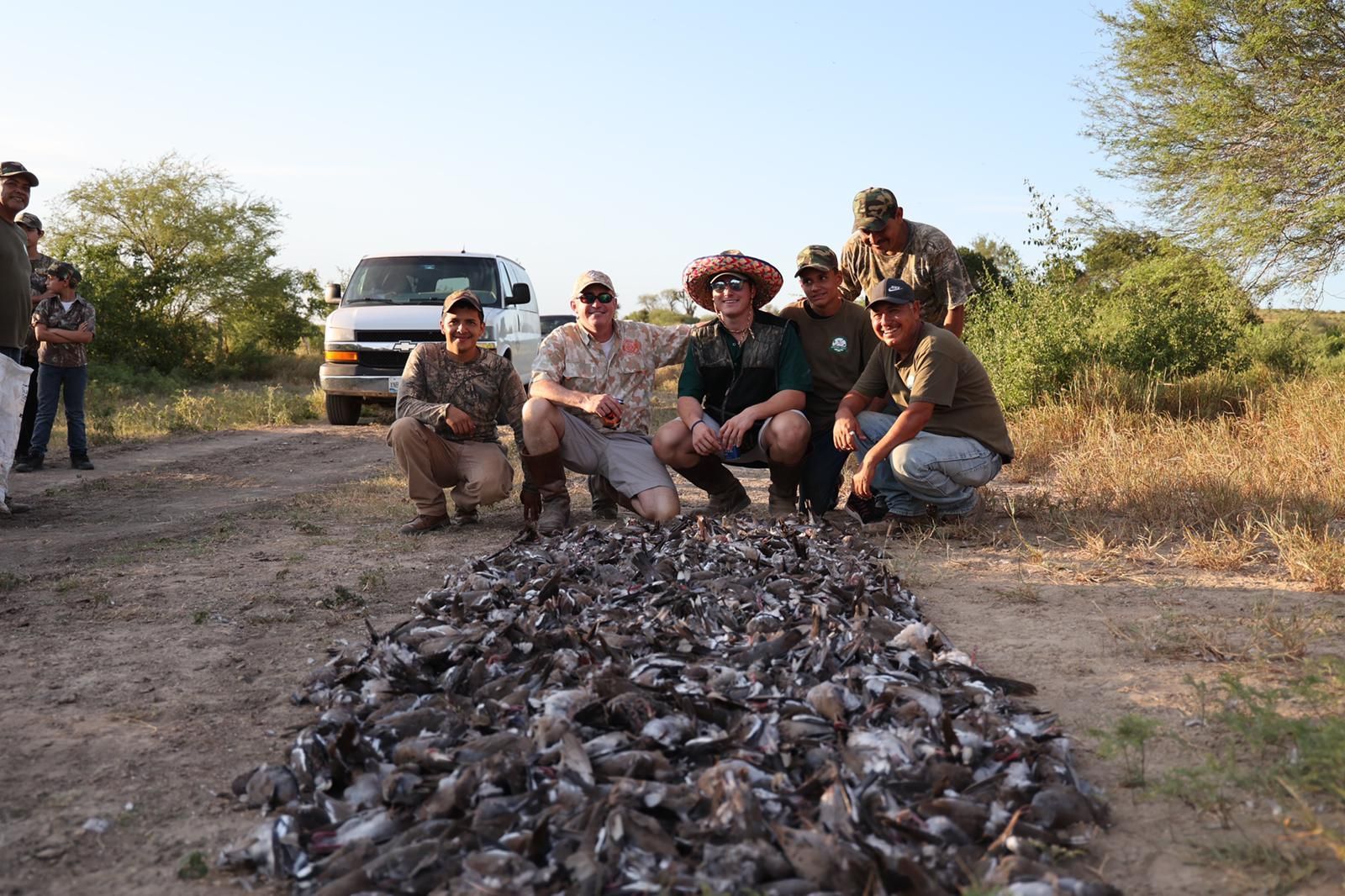Hunters kneel beside a large pile of dead birds on a dirt road, with a truck and dry brush behind them.