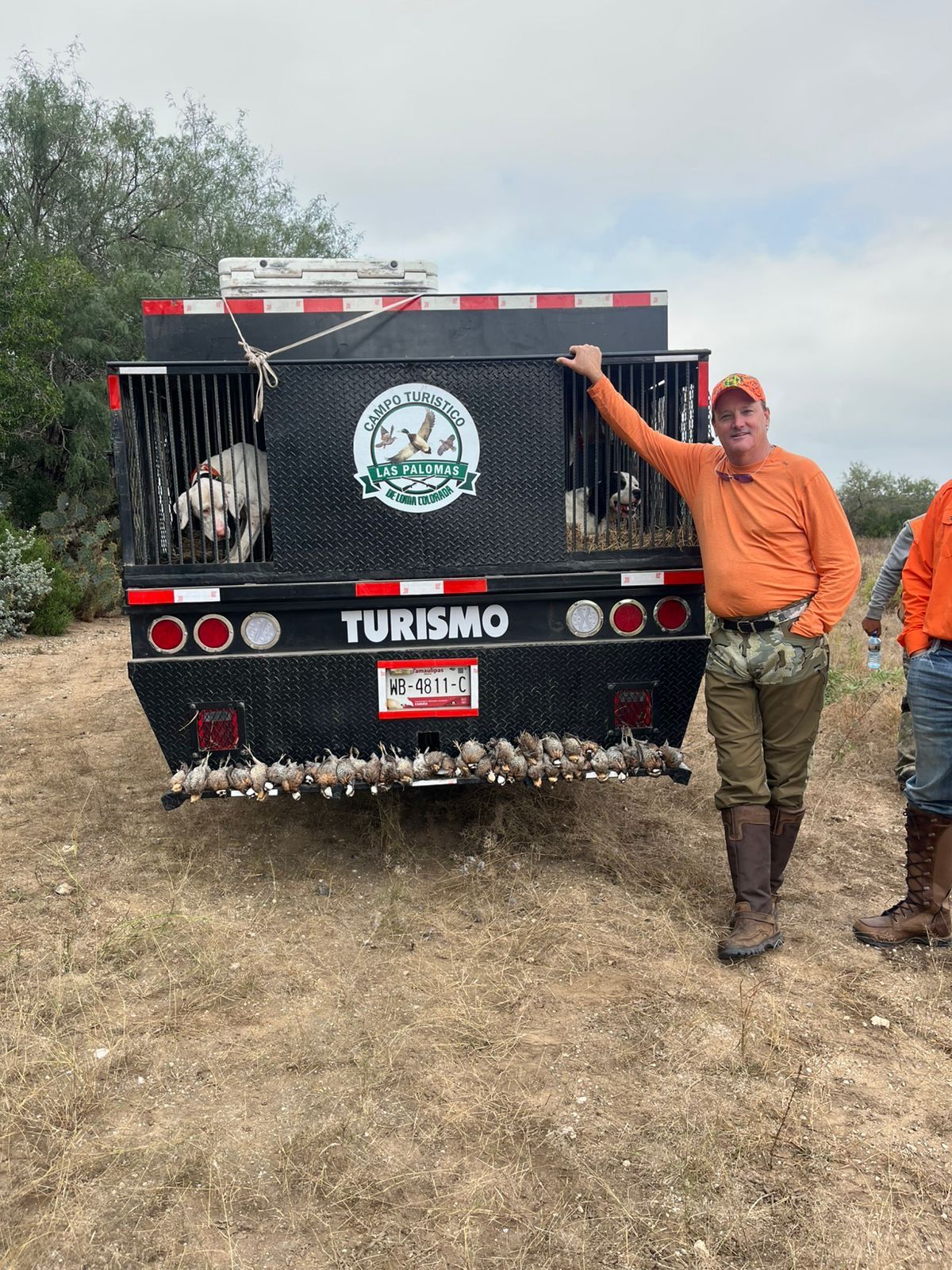 Man in orange shirt and camo boots beside a black Turismo truck in a grassy field