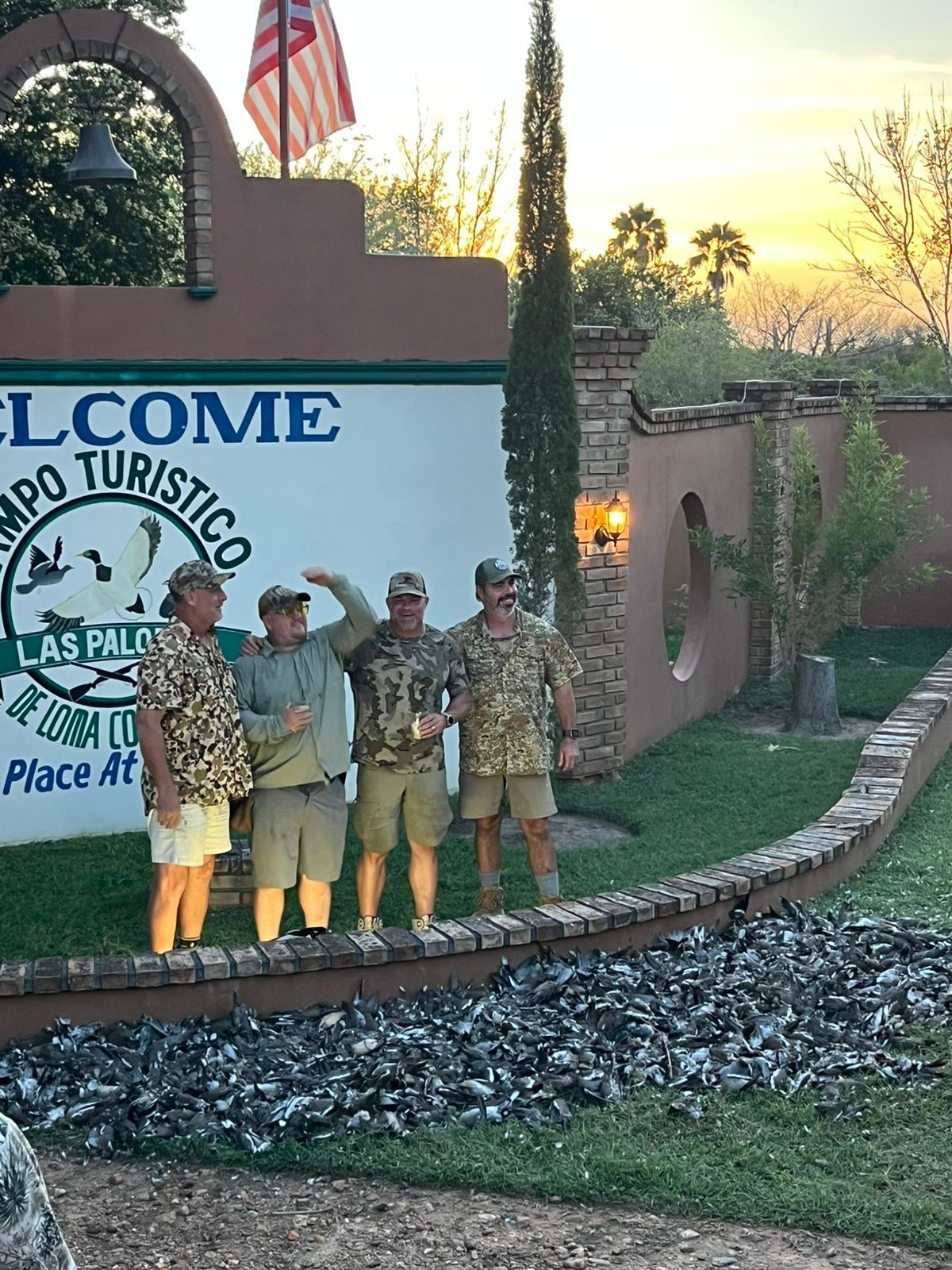 Five people posing at a lit tourist welcome sign at sunset, with palm trees and a landscaped border.
