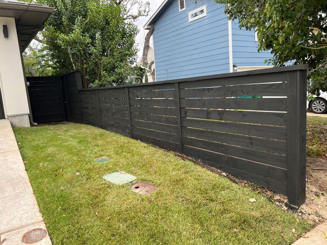 A black wooden fence surrounds a lush green yard in front of a blue house.