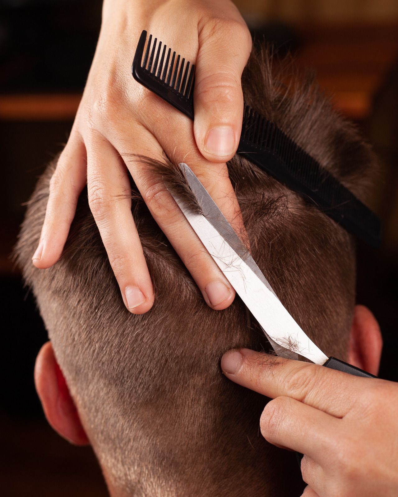 Hairdresser cutting a person's hair with scissors, using a comb to guide. The person is facing away.
