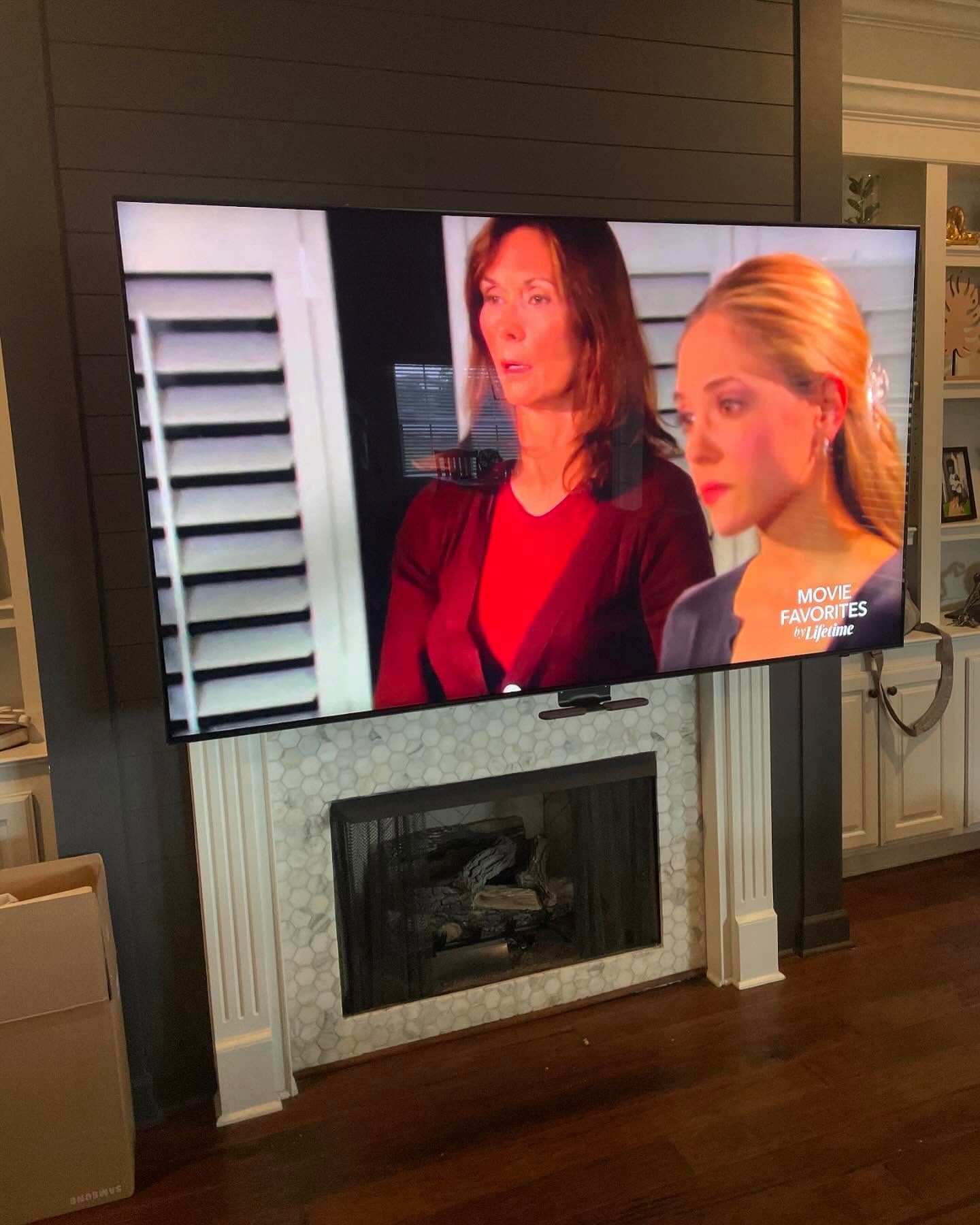 Television above a fireplace, showing two women on screen, mounted on a white and gray brick mantel. Dark wood floor.