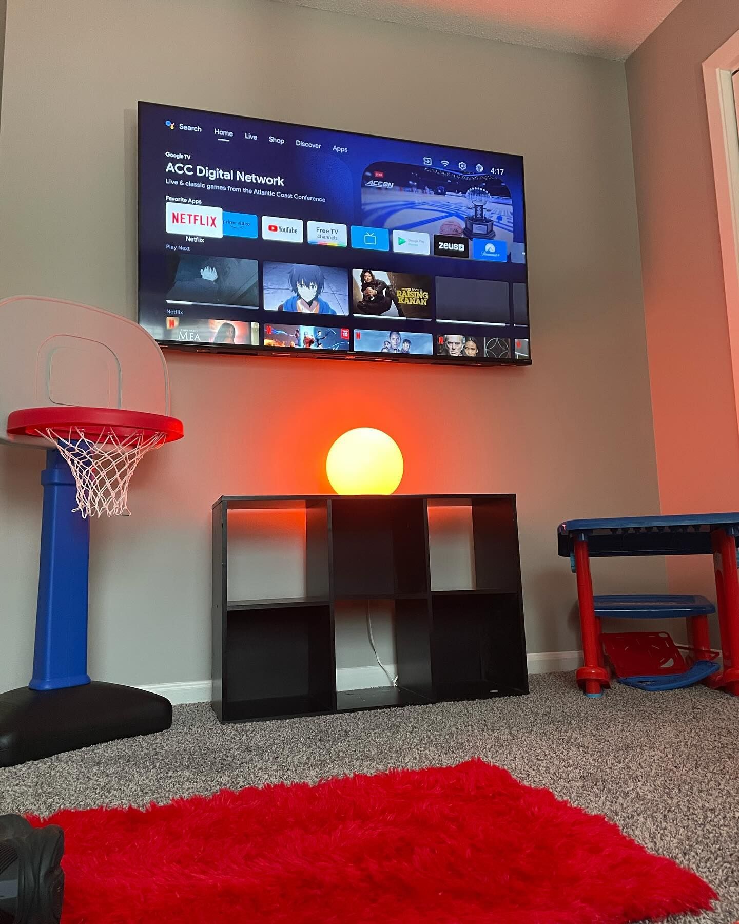 Room with a mounted TV, basketball hoop, cube shelf, and red shag rug. A glowing orange lamp sits on the shelf.
