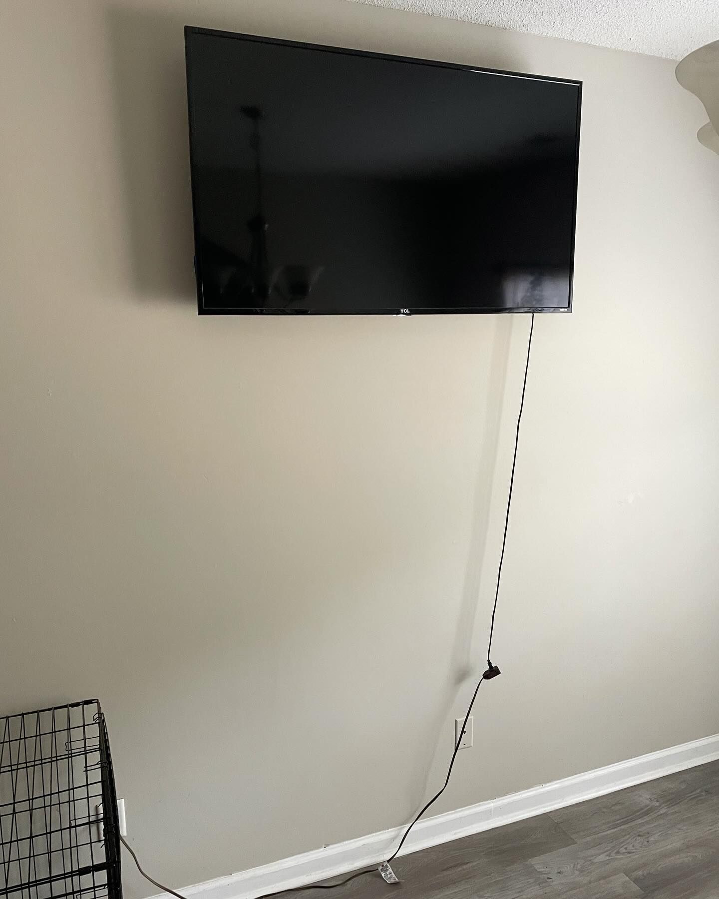 A large TV mounted on a light beige wall with a dangling power cord and a small object at the bottom left.