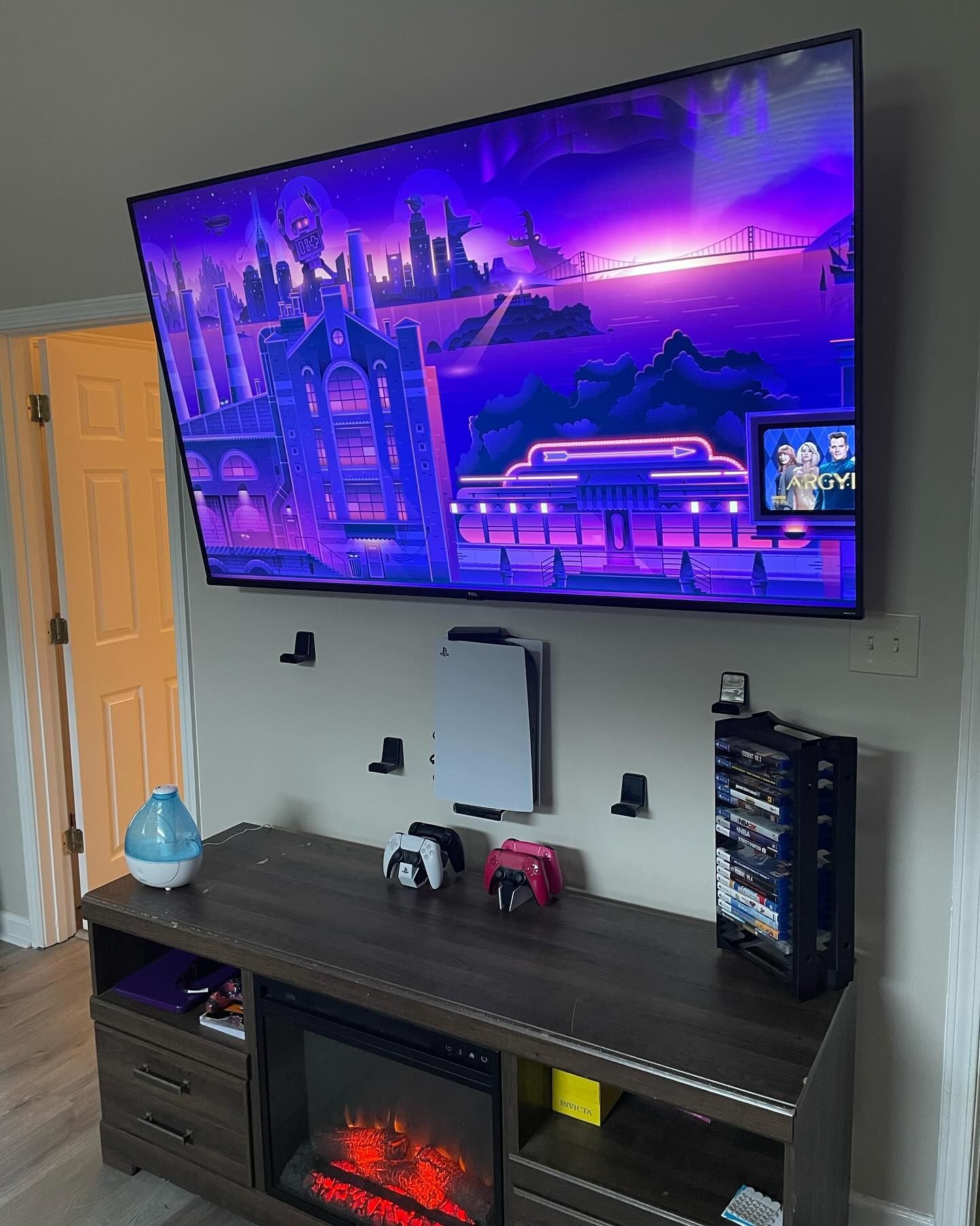 Wall-mounted TV displaying a purple cityscape, above a console with a PS5, controllers, and storage, and a fireplace.