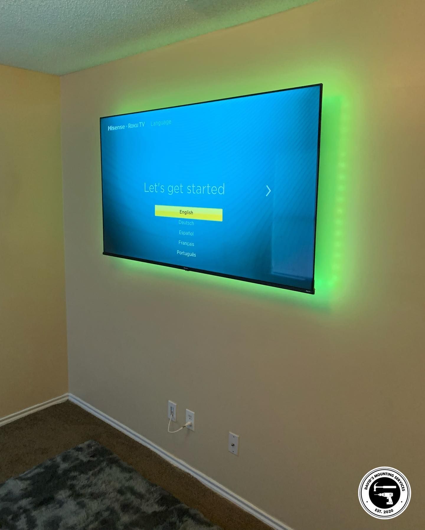 A TV mounted on a tan wall with green LED lighting around the perimeter, above a gray rug.