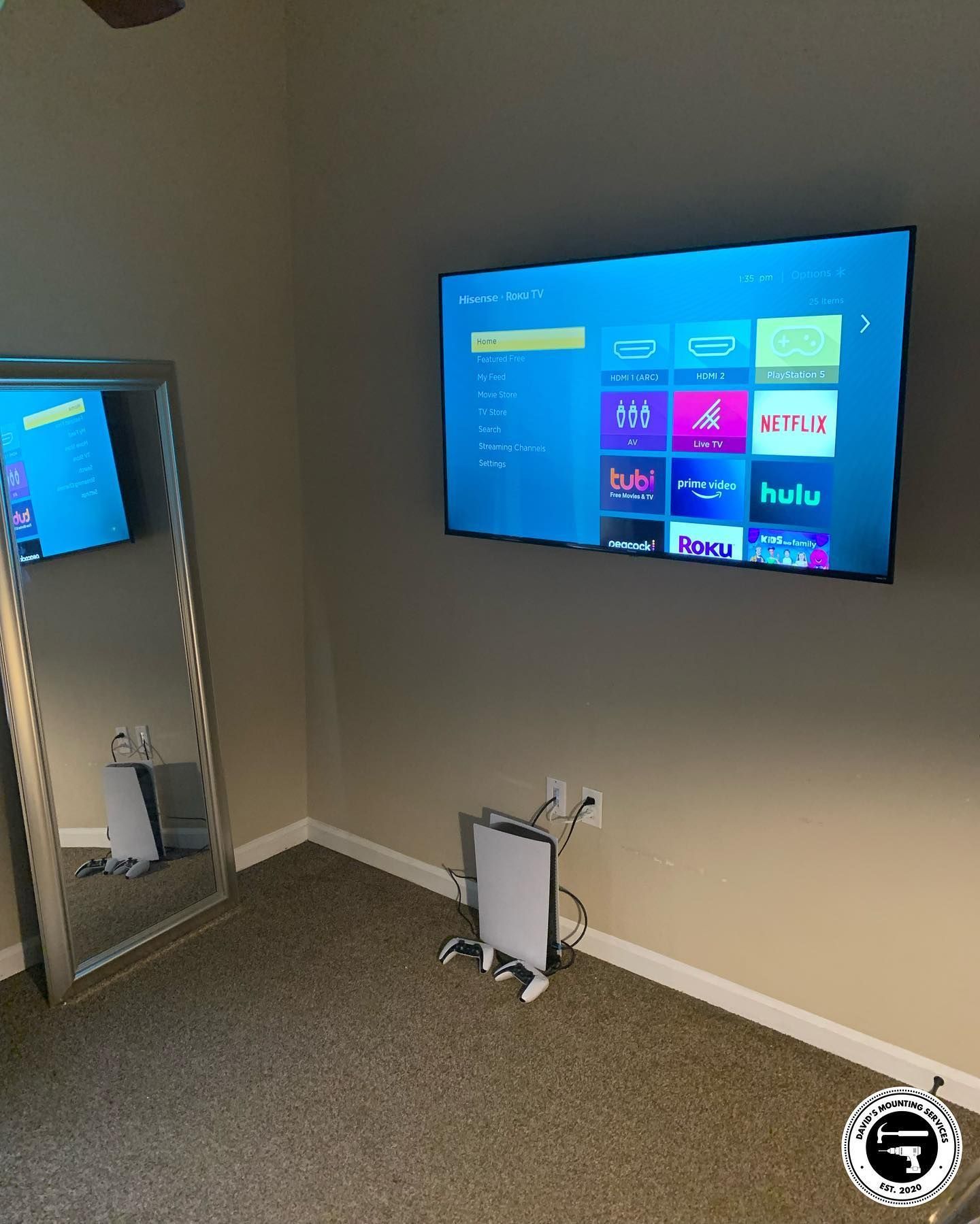 A TV mounted on a wall in a room with a full-length mirror, a game console, and cable clutter.