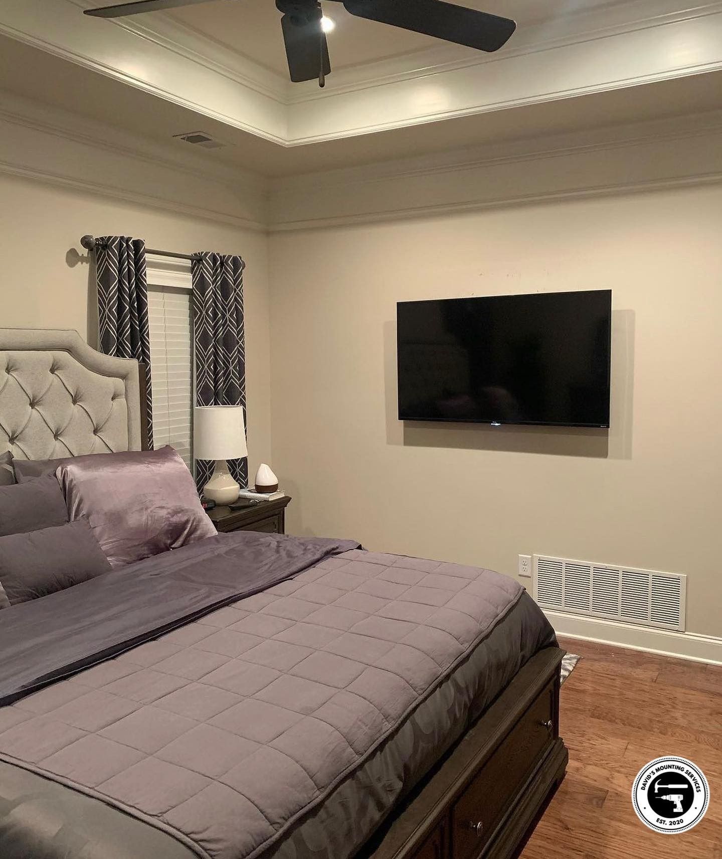 Bedroom with gray bed, wall-mounted TV, beige walls, patterned curtains, dark ceiling fan.