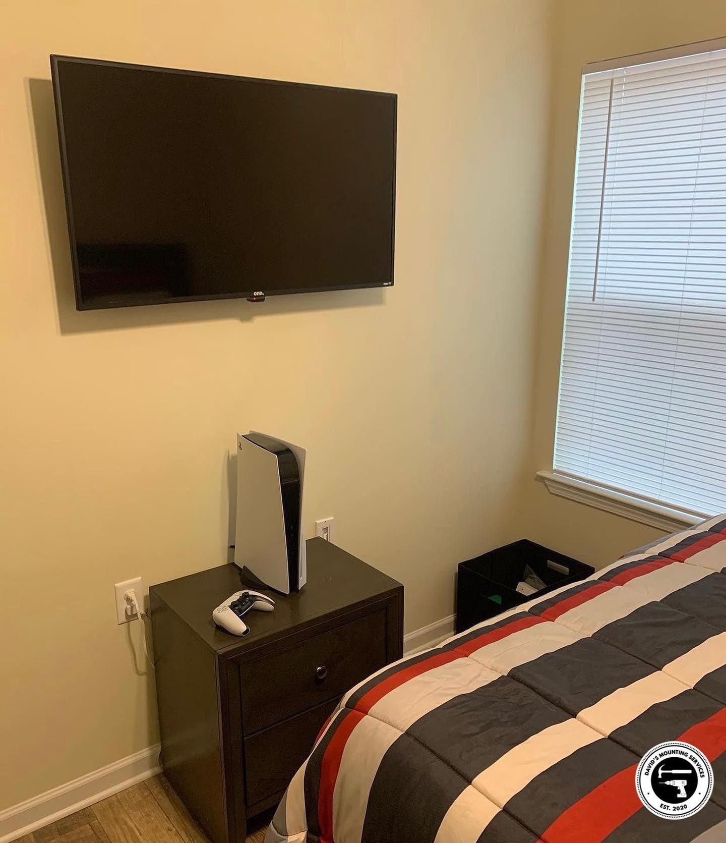 A bedroom with a TV, PS5, and a window. A bed with a striped comforter is visible.