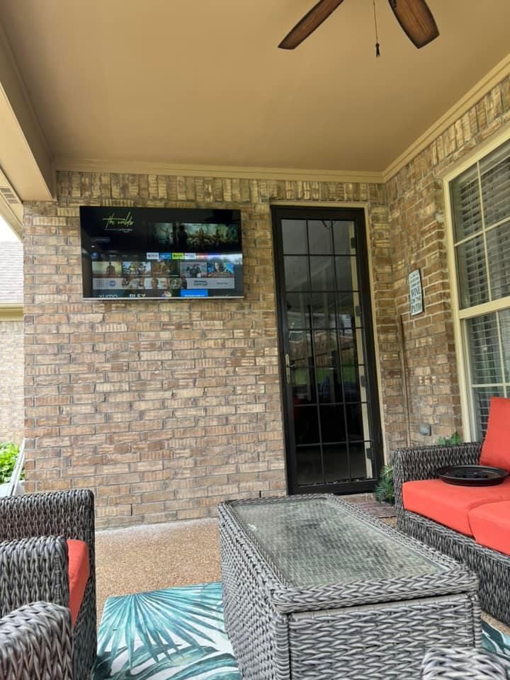 Outdoor patio with TV mounted on brick wall, glass door, seating, and ceiling fan.