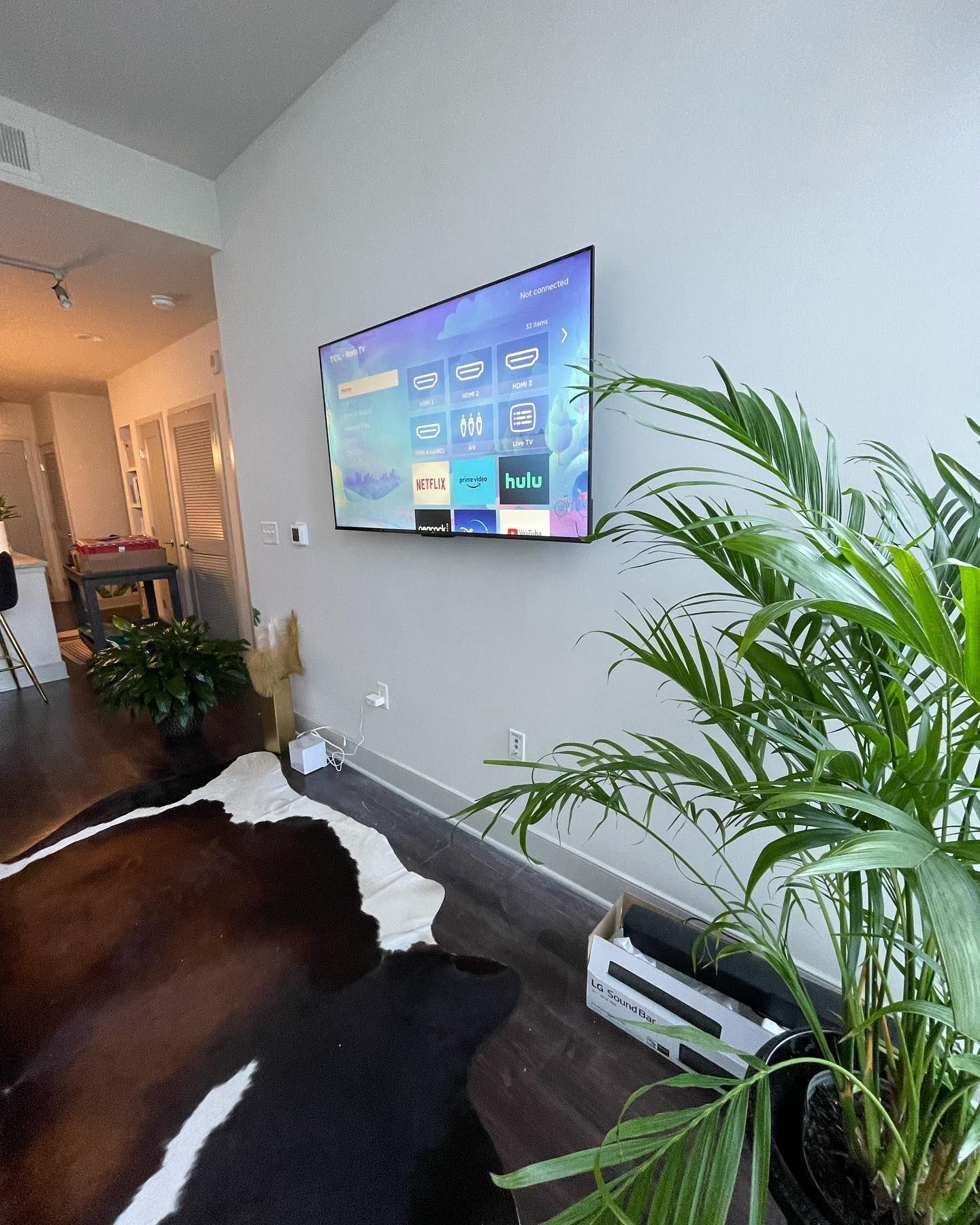 A mounted TV on a light gray wall above a cowhide rug with a plant.