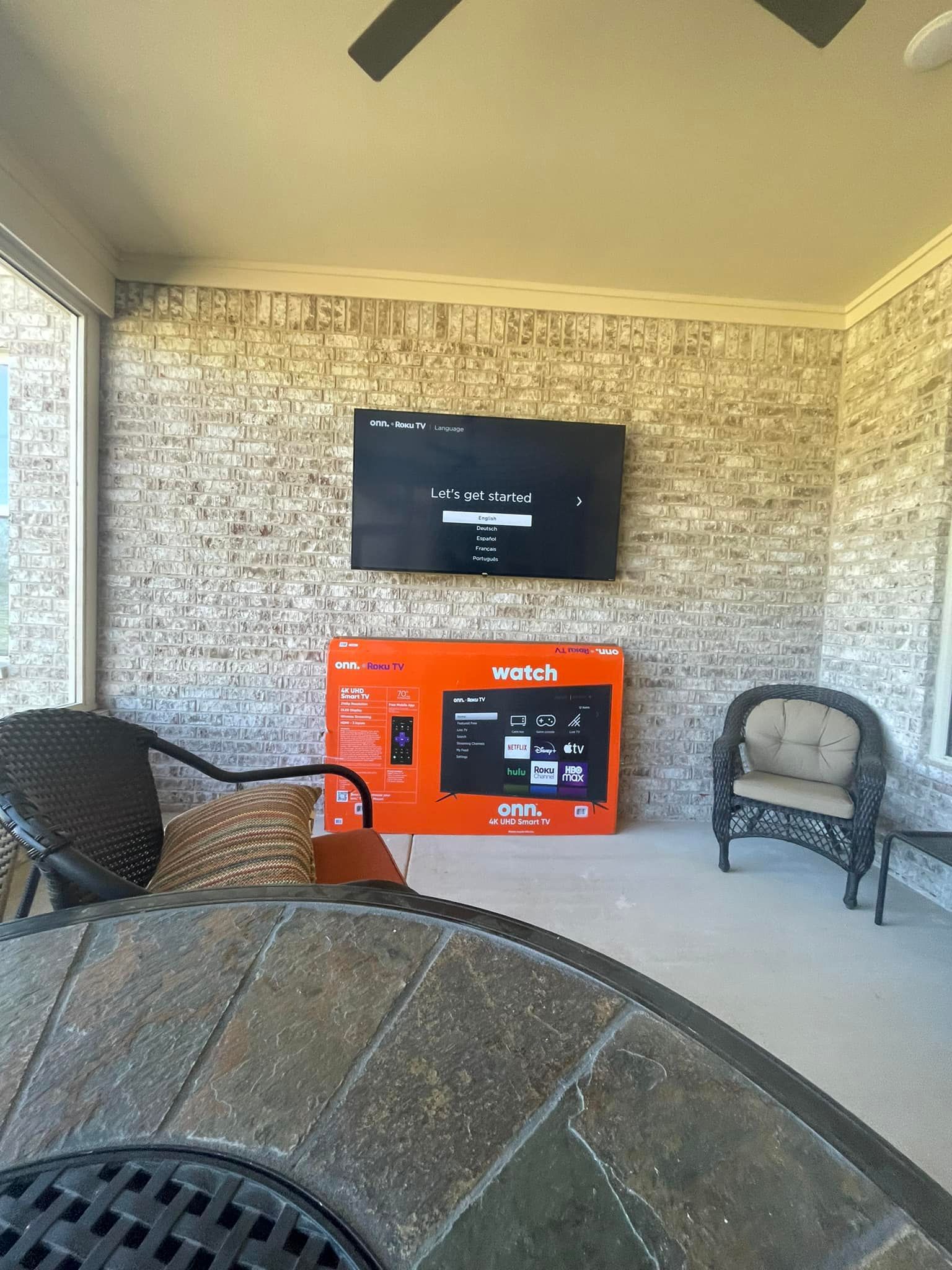 Outdoor patio with a mounted TV on brick wall, orange TV box below, and wicker furniture.