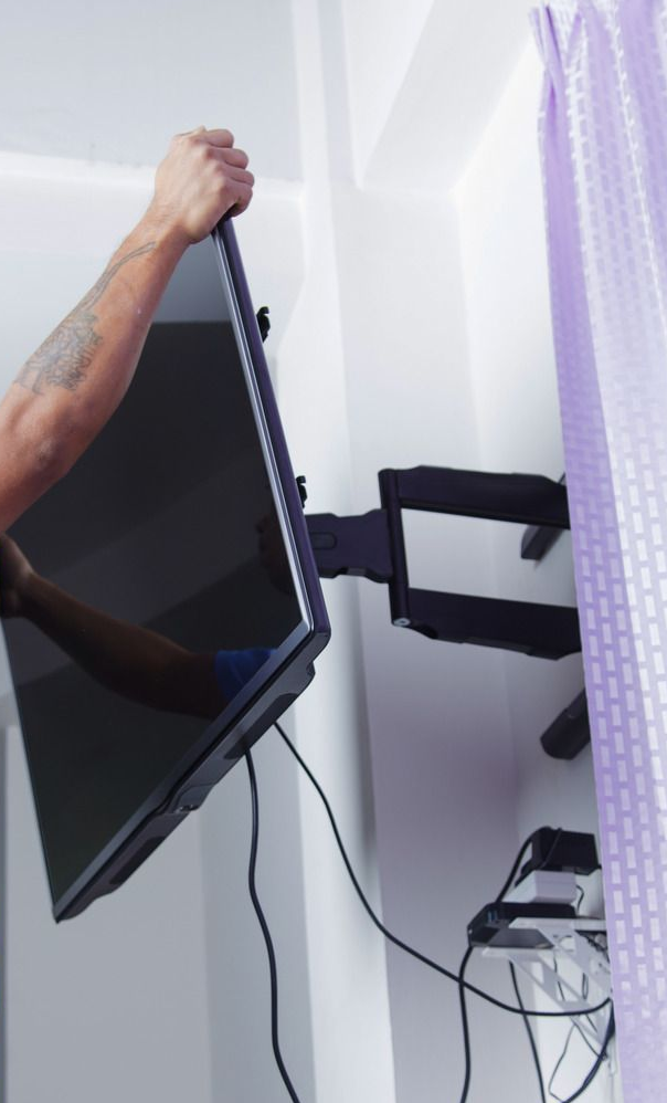 Person mounting a black flat-screen TV on a wall-mounted articulating arm. Cables hang down.