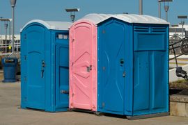 Three portable toilets—two blue and one pink—stand outdoors on a paved area.
