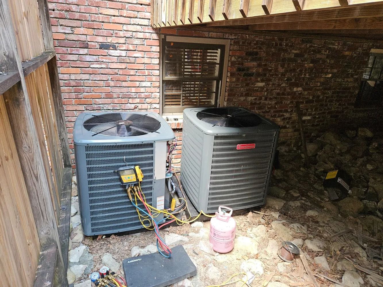 Two outdoor air conditioning units beside a brick wall, with hoses and a propane tank on the ground.