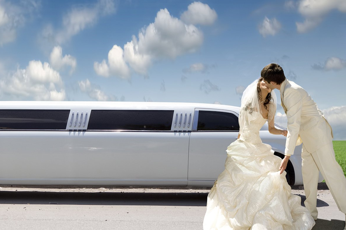 A bride and groom kissing in front of a white limousine.