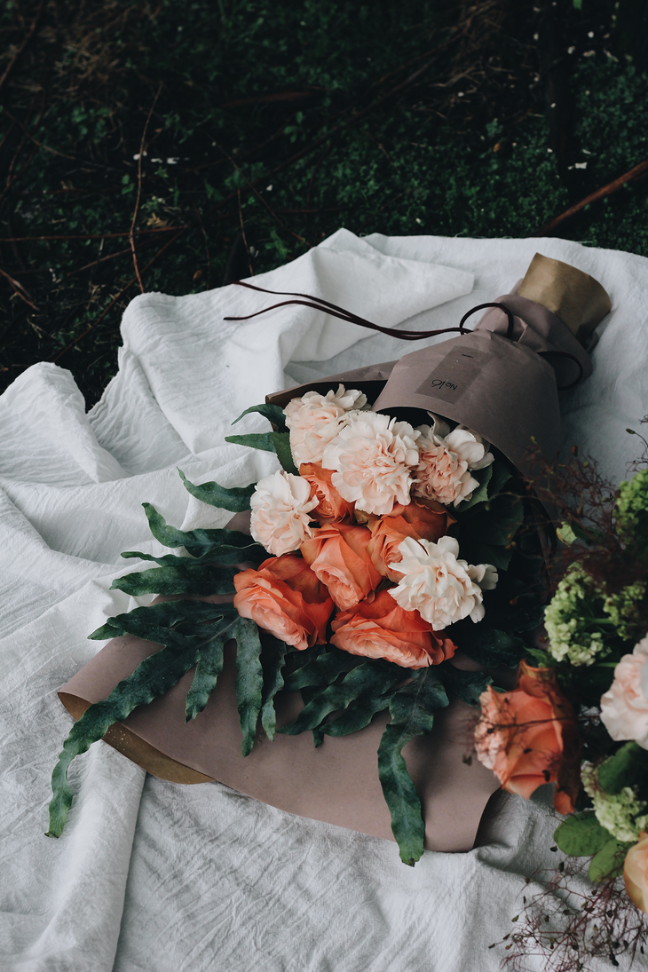 Bouquet of peach and cream flowers wrapped in brown paper, resting on white cloth against a grassy background.