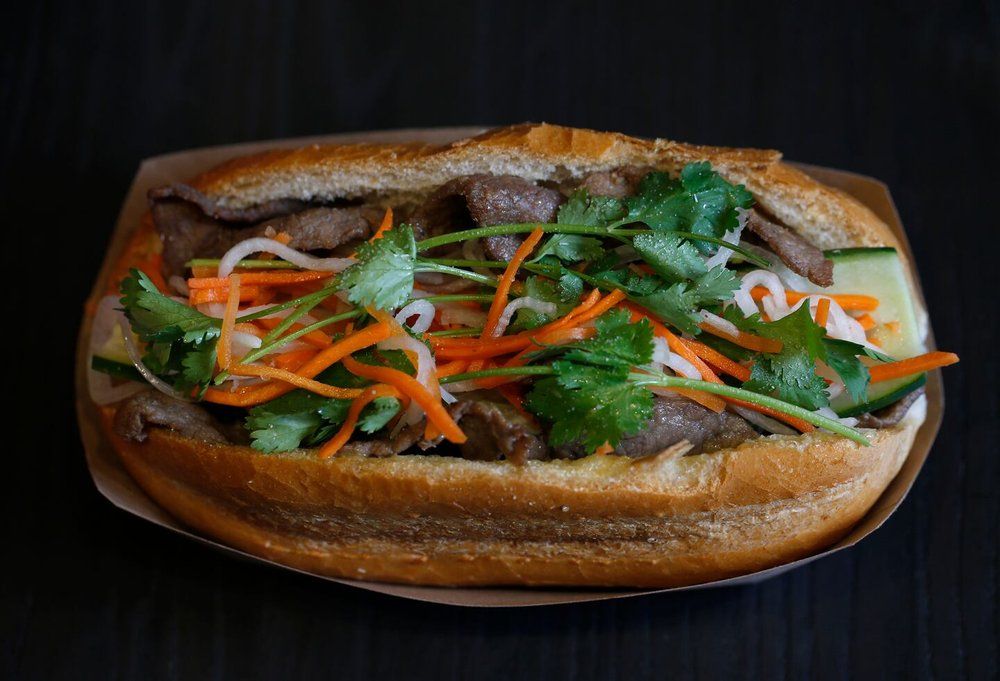 A Vietnamese banh mi sandwich with meat , carrots , cucumbers and cilantro on a bun on a paper plate.