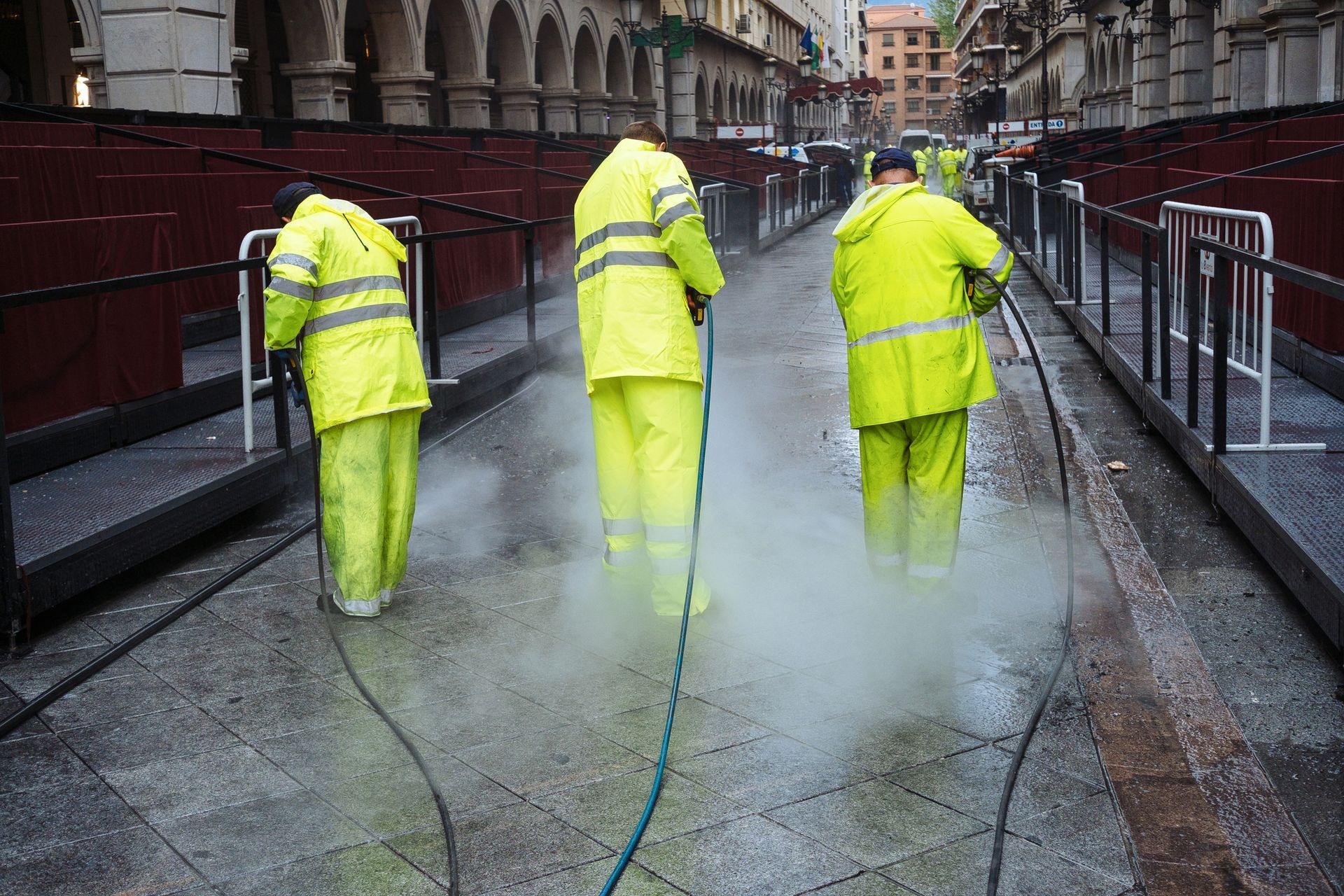 Three men in yellow safety suits are cleaning a sidewalk with a hose.