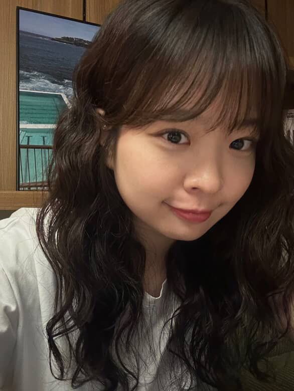 Woman with long wavy dark hair smiling indoors, wearing a light sweater.