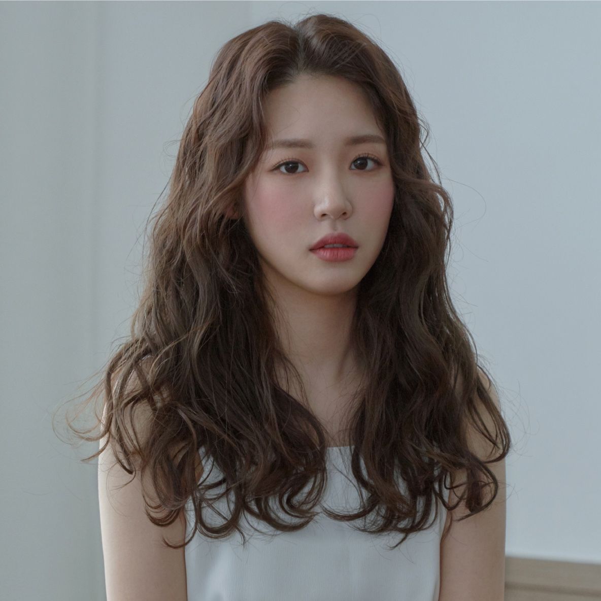 Portrait of a woman with long wavy brown hair in a white top, standing against a light wall.