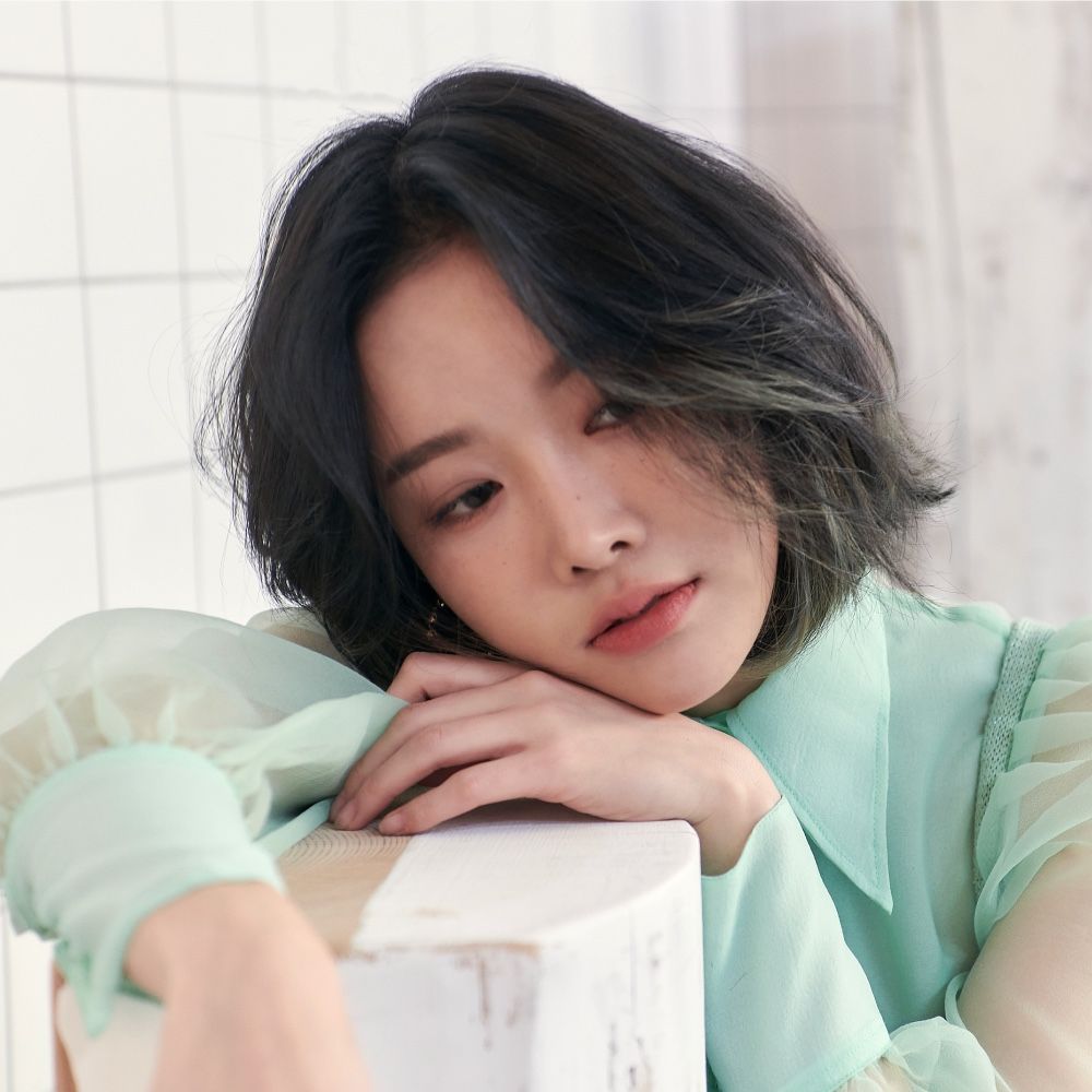 Person with short dark hair resting on a white block, wearing a mint green top in a soft-lit interior.