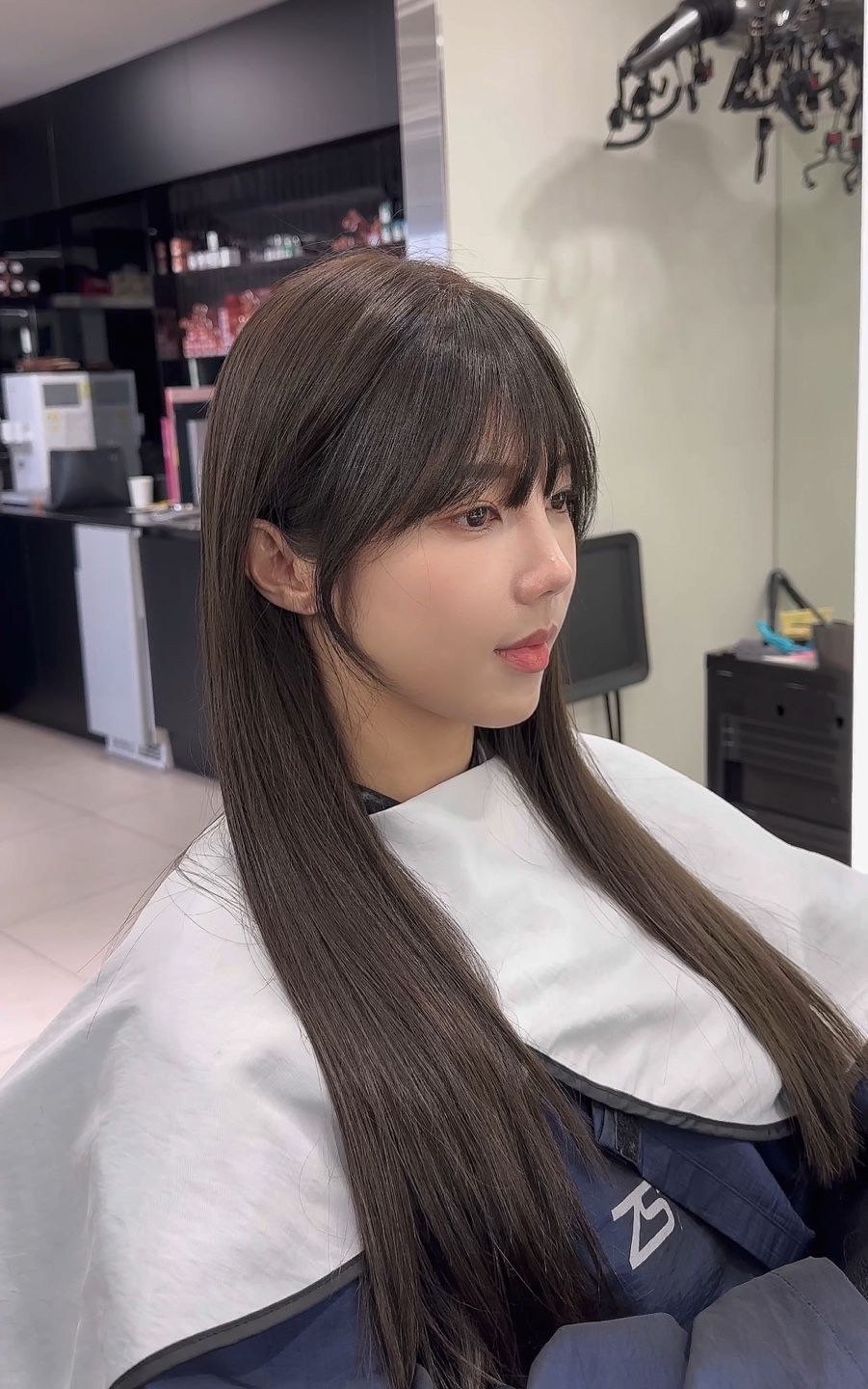 Person with long braided hair seated in a salon chair, wearing a white cape, looking to the side.