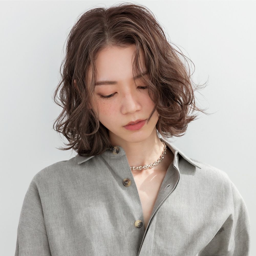 Person with wavy short hair in a gray shirt, looking down against a plain light background