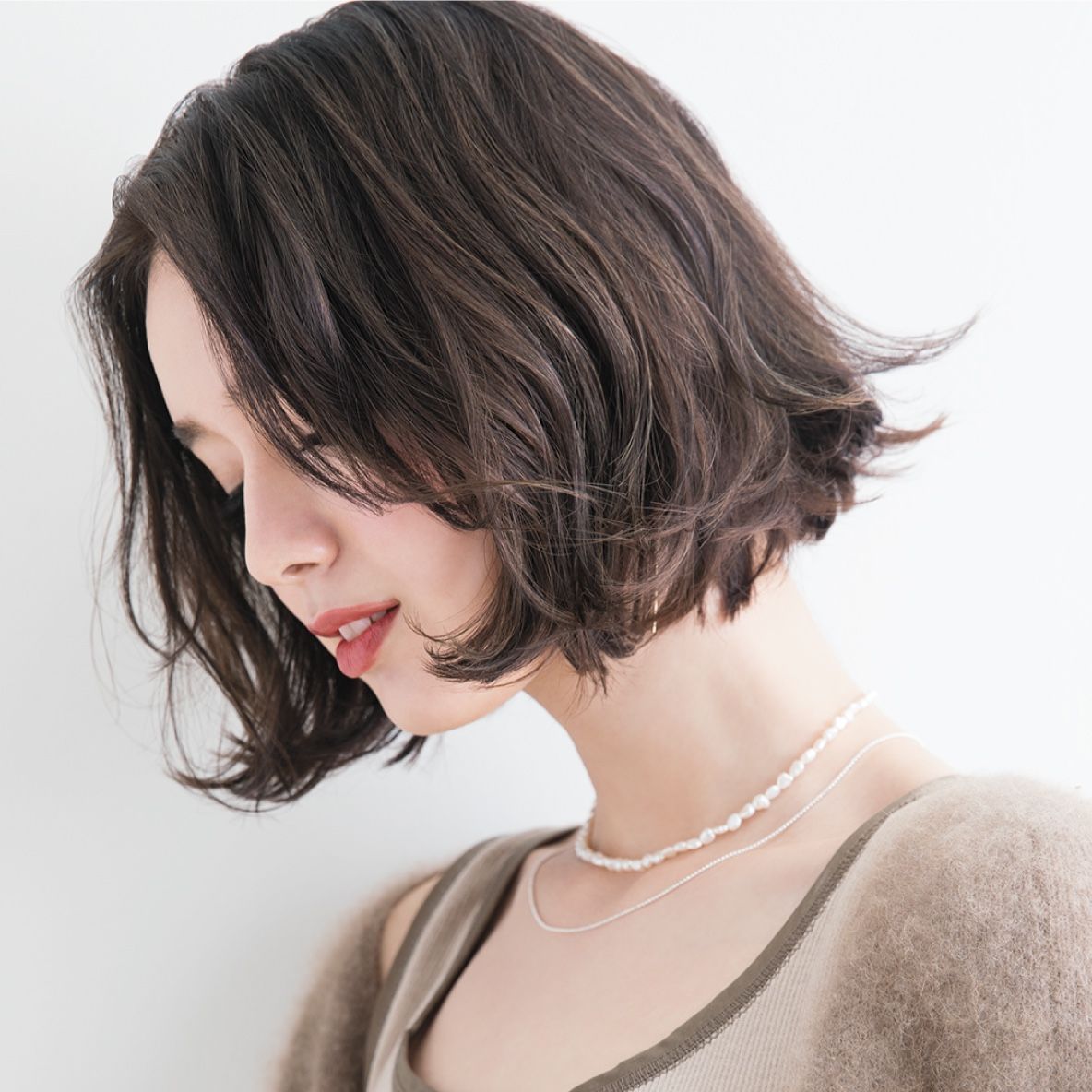 Profile of a woman with short wavy dark hair, looking down in a beige top and pearl necklace