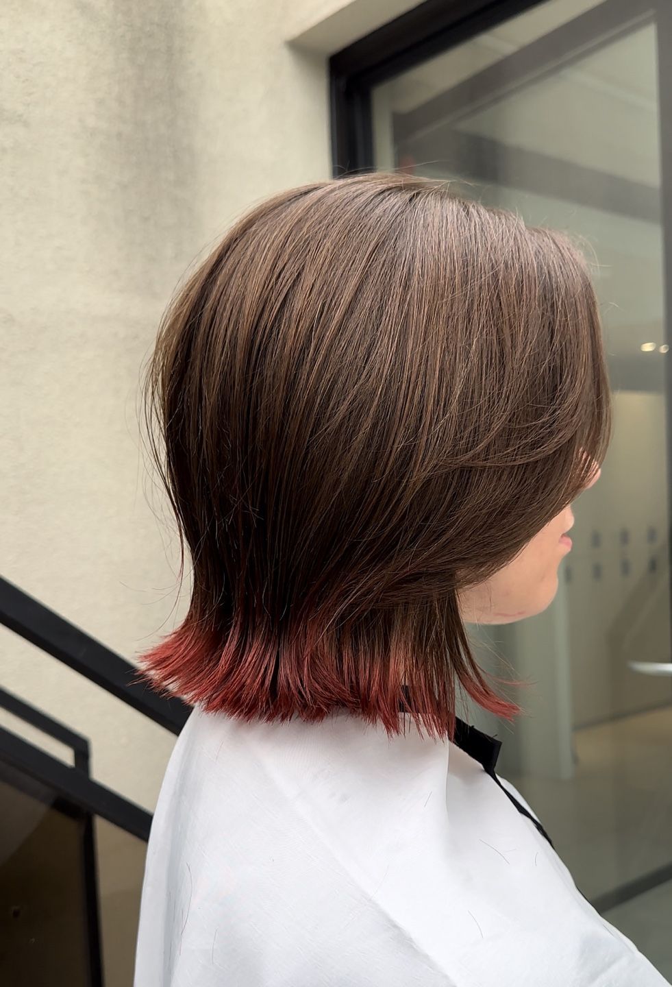 Side view of a layered brown bob with red-dyed ends against an outdoor building backdrop