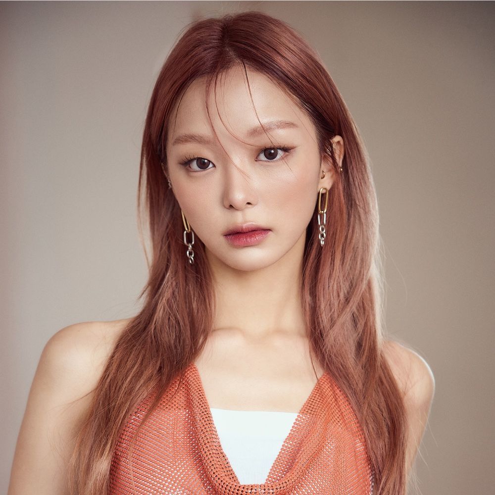 Portrait of a person with auburn hair in an orange top, looking at the camera against a plain beige background