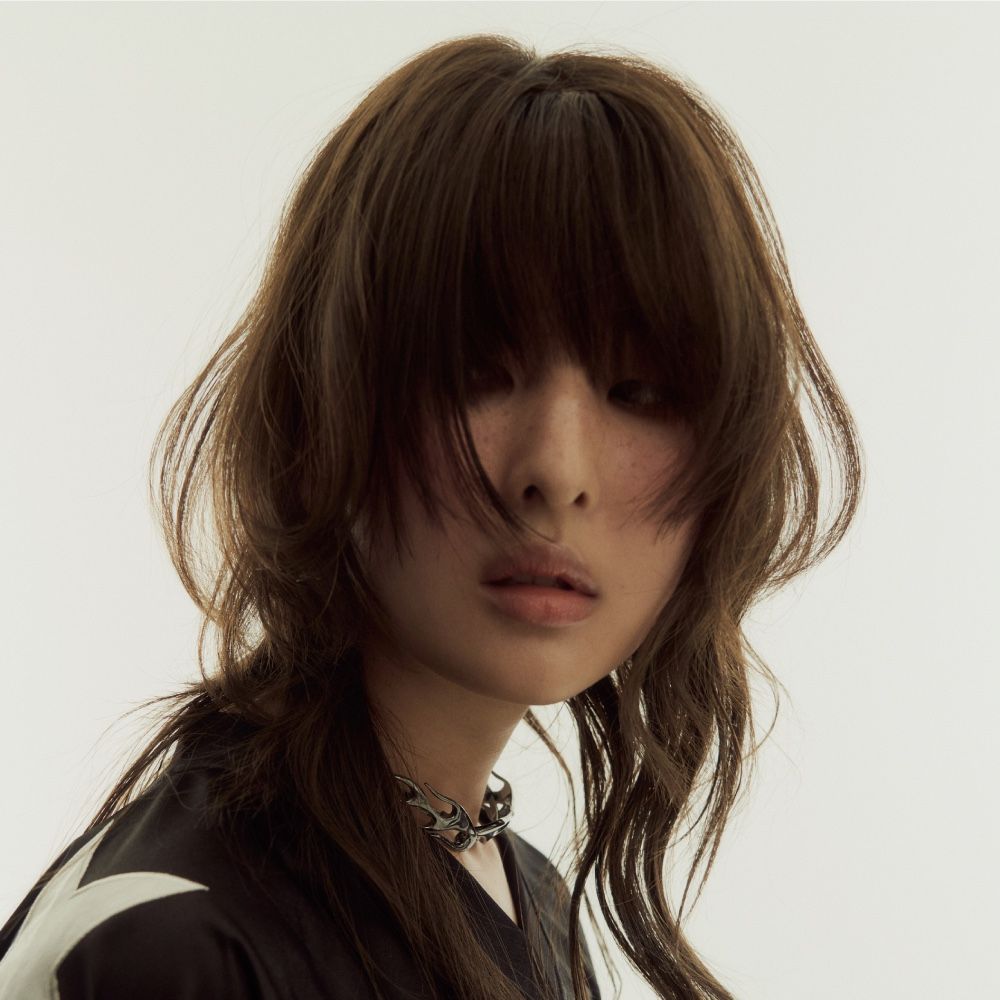Close-up portrait of a person with shaggy brown hair and a neutral expression on a plain background