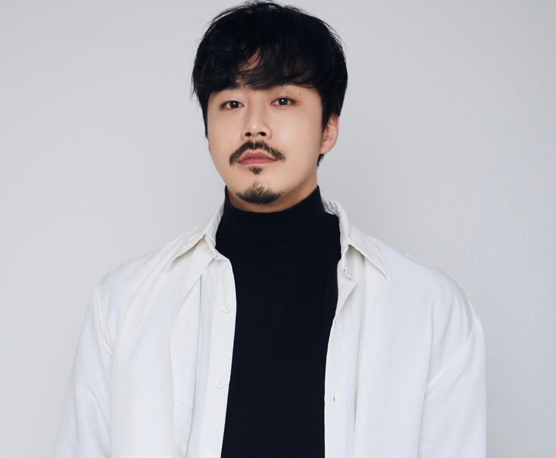 Portrait of a person with black hair and mustache, wearing a white shirt over a black turtleneck against a gray background