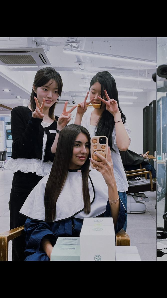 Three people posing in a salon mirror selfie, two standing behind one seated, all making peace signs.
