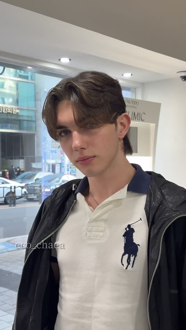 Young person in a black jacket and white polo shirt standing indoors near a window